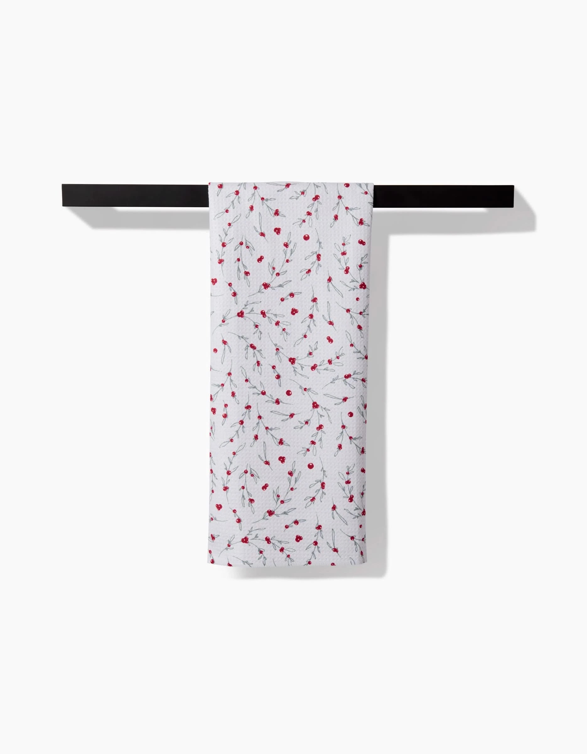 The Ashley Celena Holiday Sprigs tea towel, crafted from recycled materials, features a charming pattern of small red berries and green leaves. It hangs neatly over a black bar against a plain white background for an eco-friendly touch.