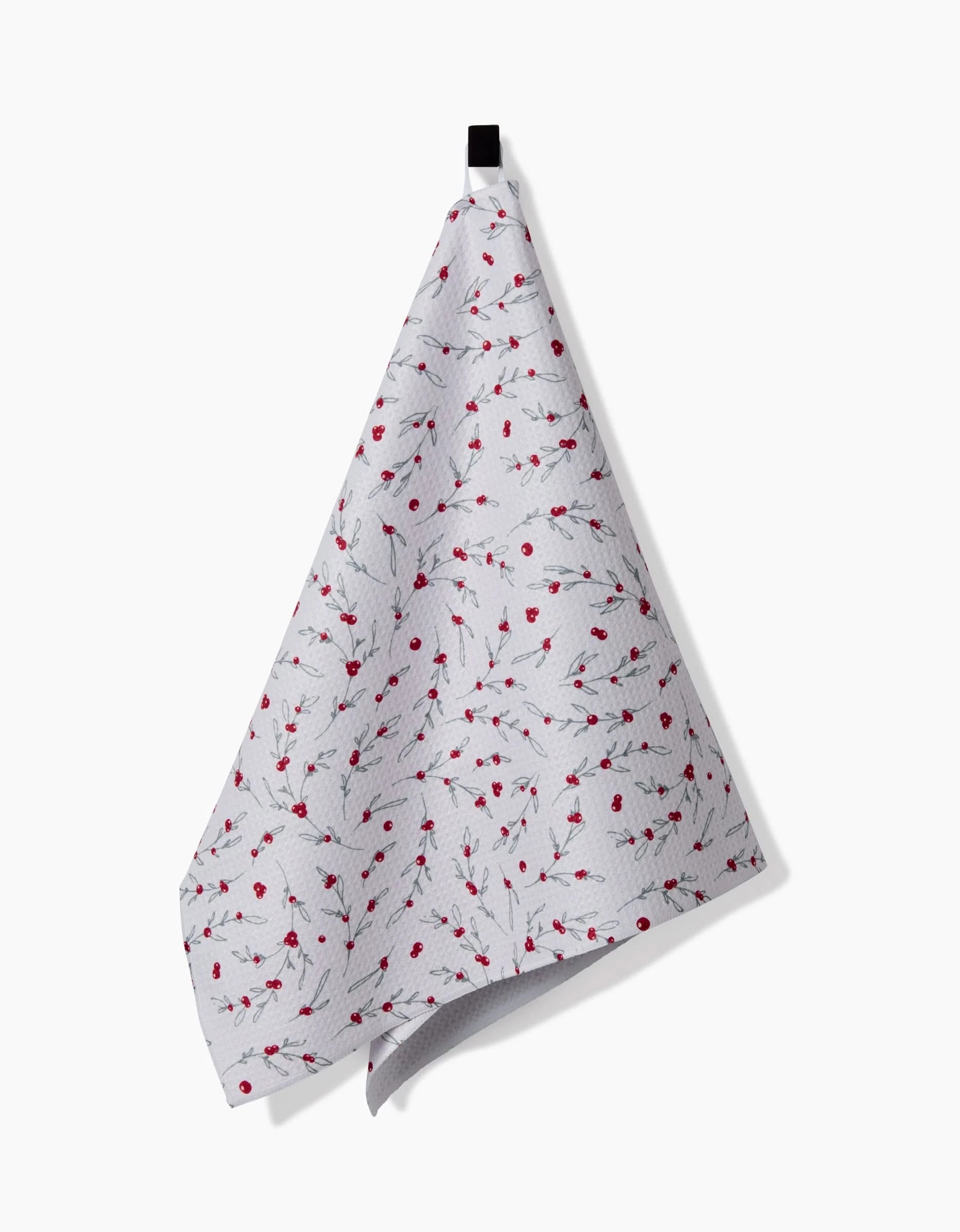 The Holiday Sprigs tea towel by Ashley Celena features a white base with subtle leaves and small red flowers, made from recycled materials. It hangs from a black loop against a plain white background.