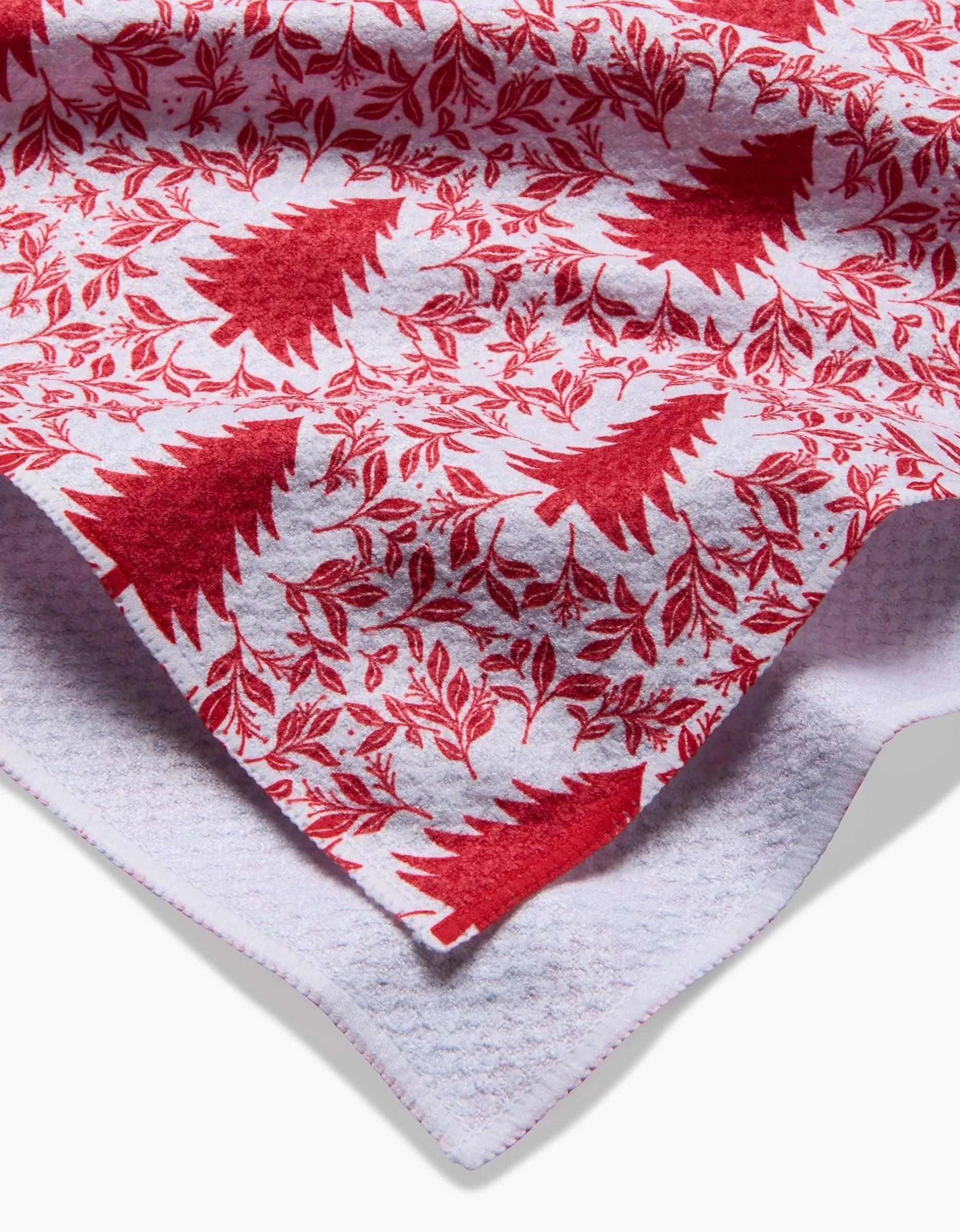 A close-up of the Charlie Rowan Designs Holiday Tree towel, featuring ultra-absorbent white fabric with red tree and leafy patterns, layered corners, and a textured underside.