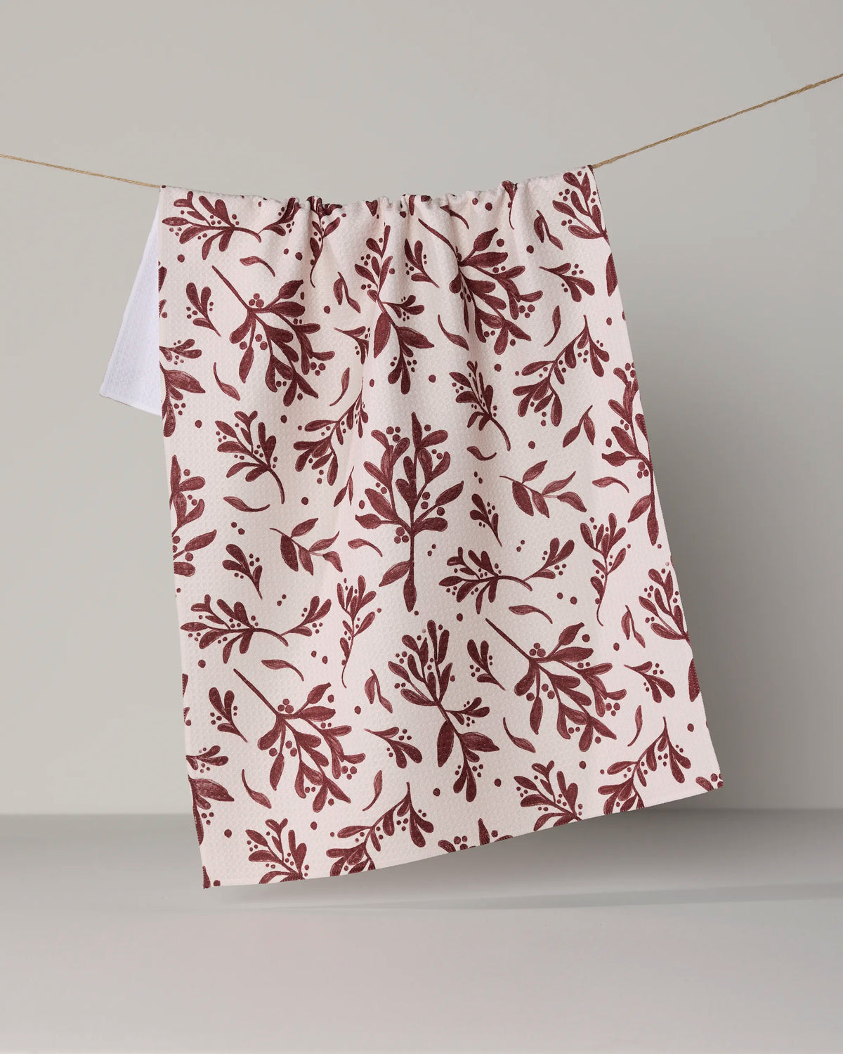 A Geometry “Holly Folly” tea towel, featuring a maroon botanical leaf pattern on white, hangs on a thin clothesline against a neutral background.