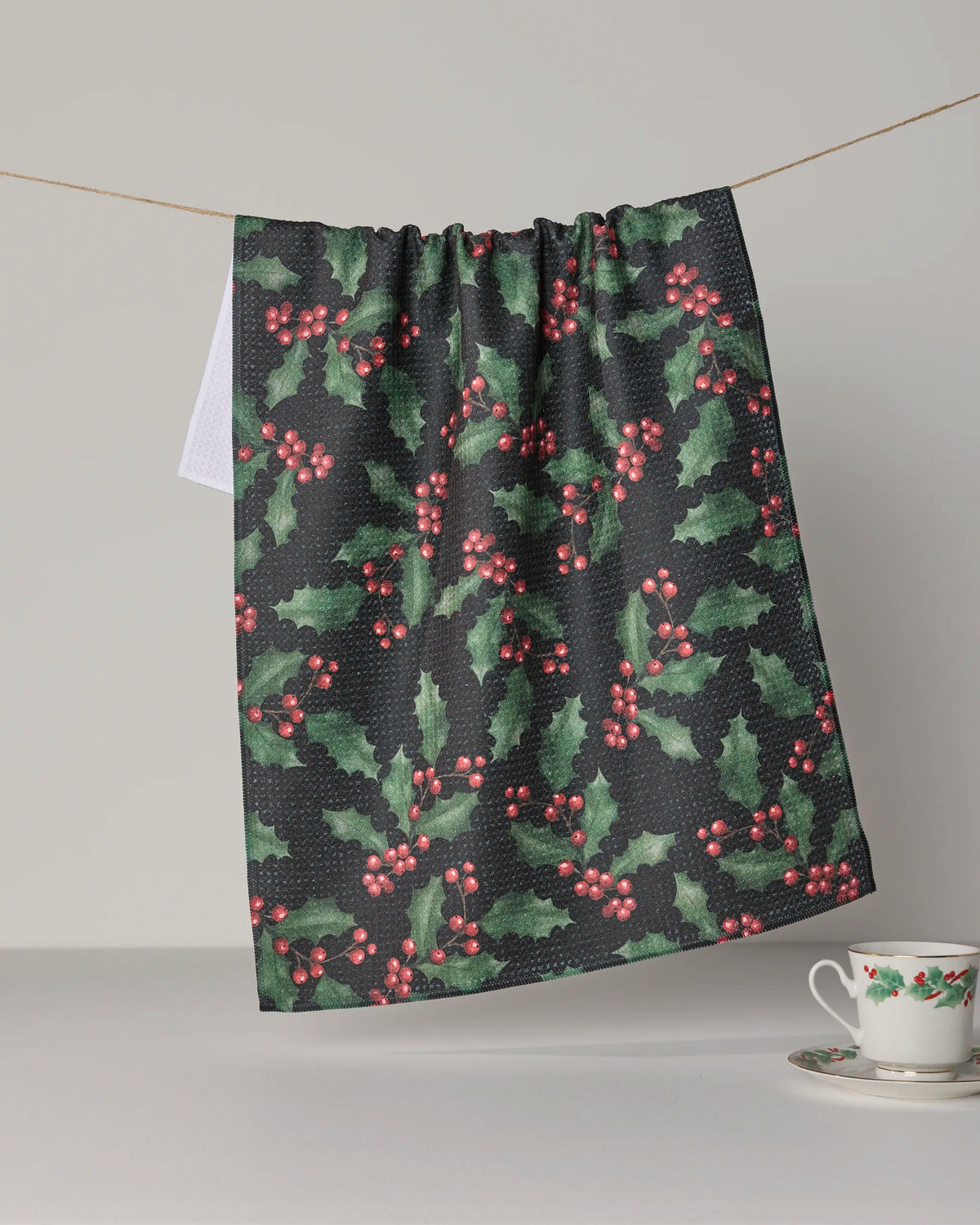 A kitchen tea towel from Geometry’s “Holly Night” collection, featuring a holly leaf and red berry pattern and crafted from recycled materials, hangs on a line. Nearby, a white teacup and saucer adorned with the matching “Holly Night” holly design rest on a light surface against a plain background.