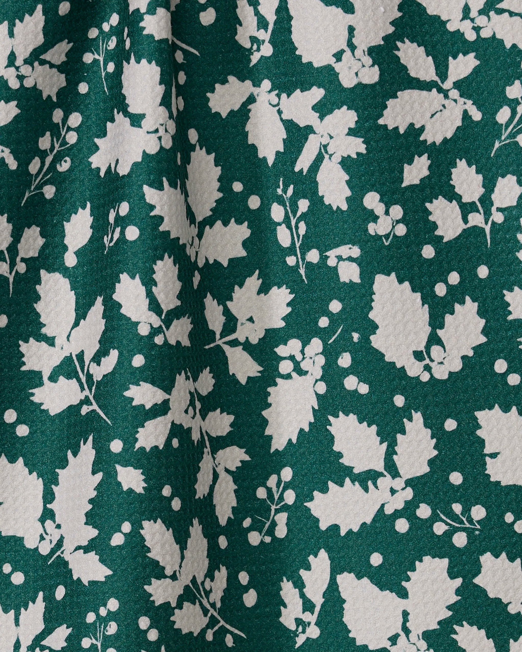 The Geometry Hollyday tea towel features ultra-absorbent green fabric with a textured surface and a white holly leaf, berry, and branch pattern for a festive botanical look.