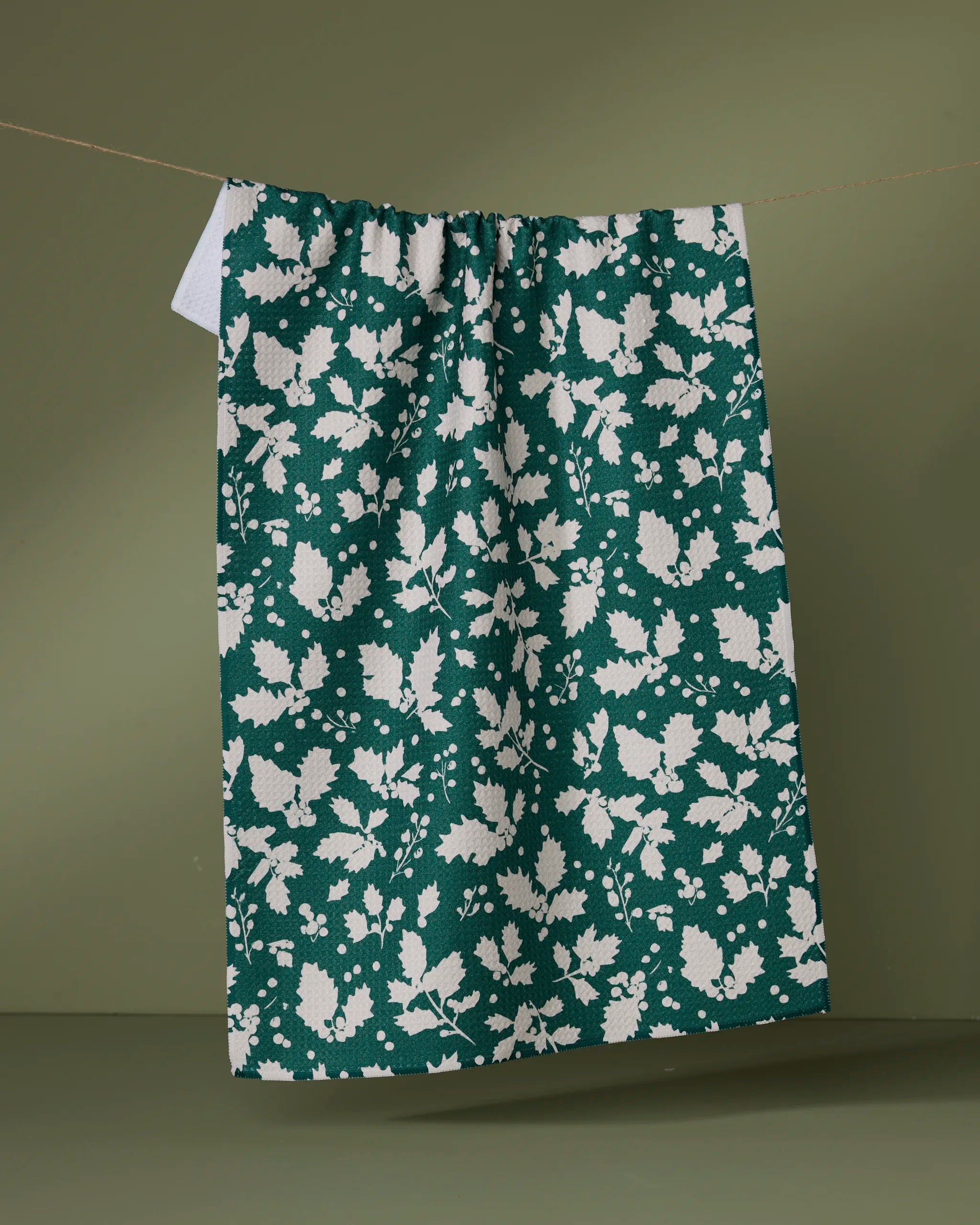 The Geometry Hollyday tea towel, ultra-absorbent and green with a white floral and leaf pattern, hangs on a clothesline with an olive green background.