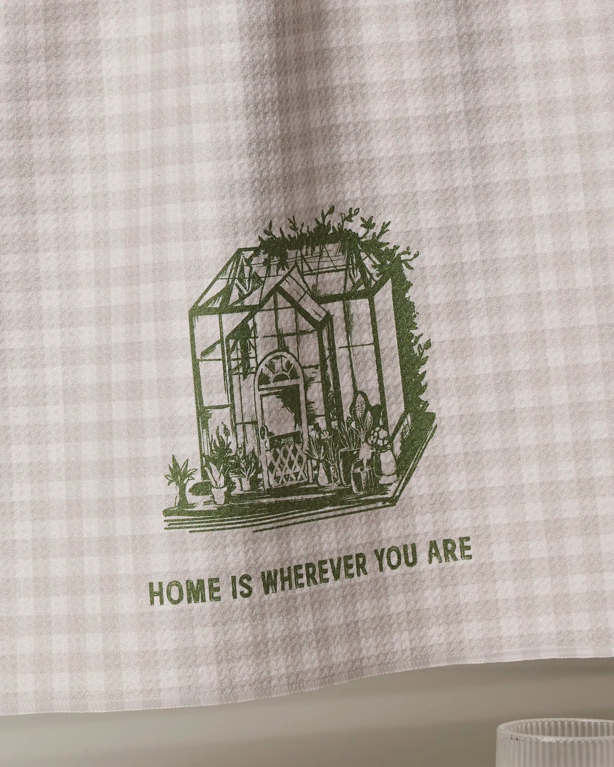 The Home is You kitchen towel by Geometry is eco-friendly and checkered, featuring a green greenhouse with potted plants and "HOME IS WHEREVER YOU ARE" printed below the illustration.