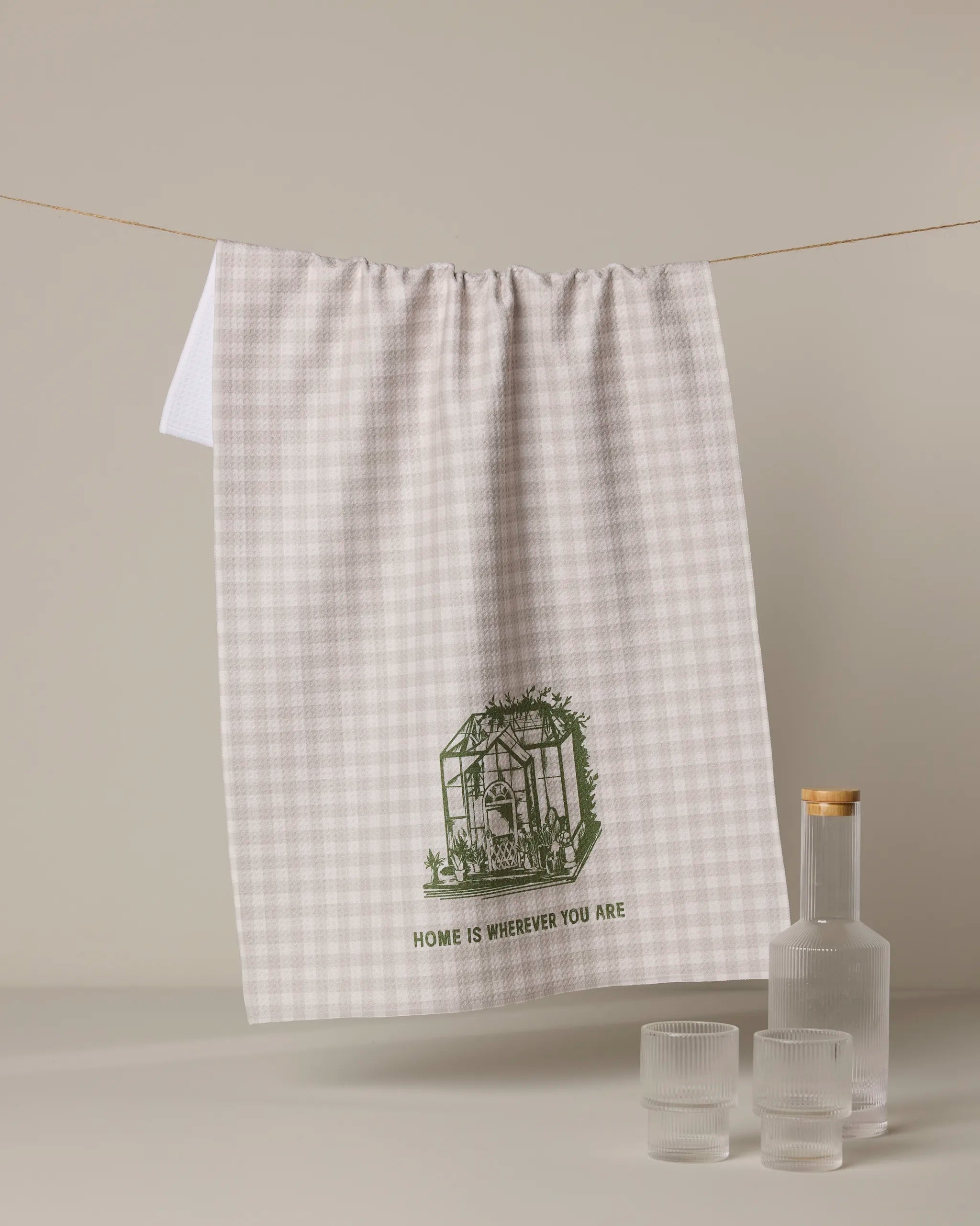 The Geometry "Home is You" dish towel, featuring a green greenhouse illustration and the words "Home is wherever you are," hangs above a table set with a clear glass bottle and two ribbed drinking glasses.