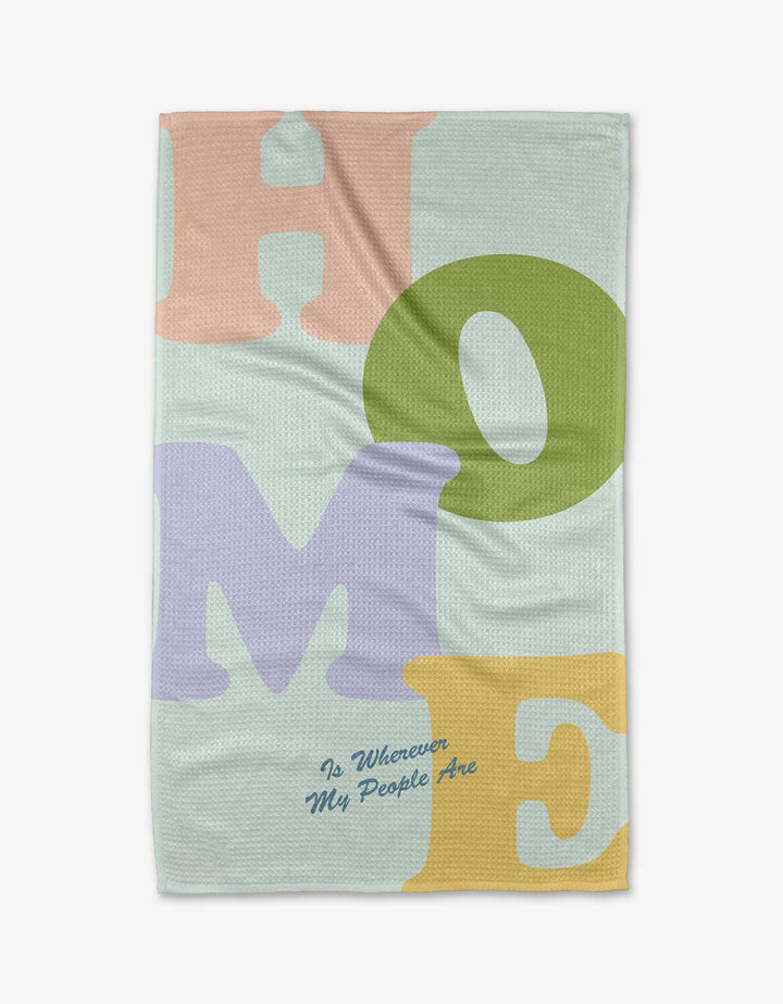 The Home tea towel by Quotes by Christie features large pastel letters spelling HOME in pink, green, purple, and yellow on recycled fabric, with “Is Wherever My People Are” in small blue cursive underneath.