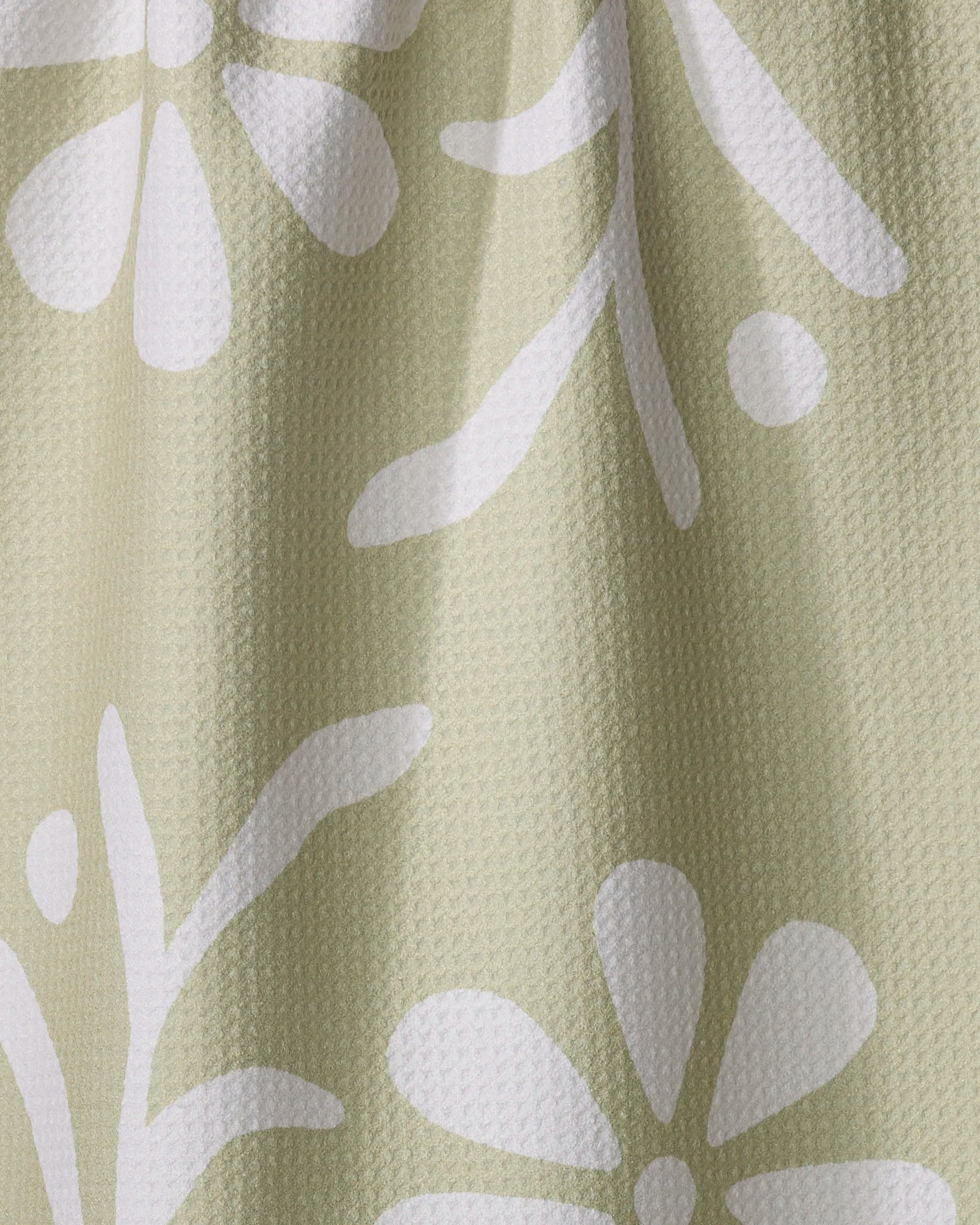A close-up of the Honey Dew tea towel by Graphics and Grain, featuring a textured light green fabric with a white abstract floral pattern and subtle folds.