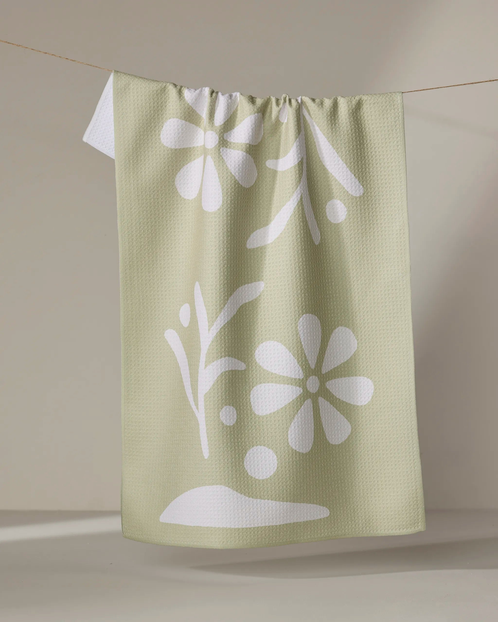 The "Honey Dew" kitchen tea towel by Graphics and Grain features light green textured fabric with white abstract floral and leaf patterns, shown hanging on a clothesline against a neutral background.