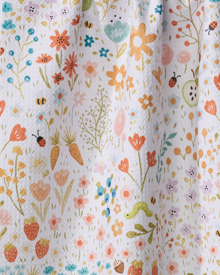 Sara Mari’s “Honeybee Heaven” is a kitchen tea towel crafted from recycled materials, featuring a spring-inspired design with flowers, strawberries, carrots, ladybugs, bees, small plants, and a green snake on a white background.