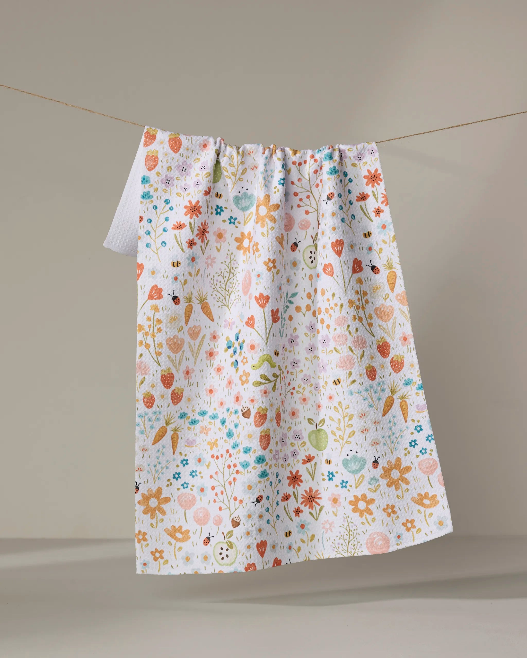 The Honeybee Heaven tea towel by Sara Mari, crafted from recycled materials, displays a colorful floral and nature-inspired design and hangs on a clothesline against a plain, neutral background.
