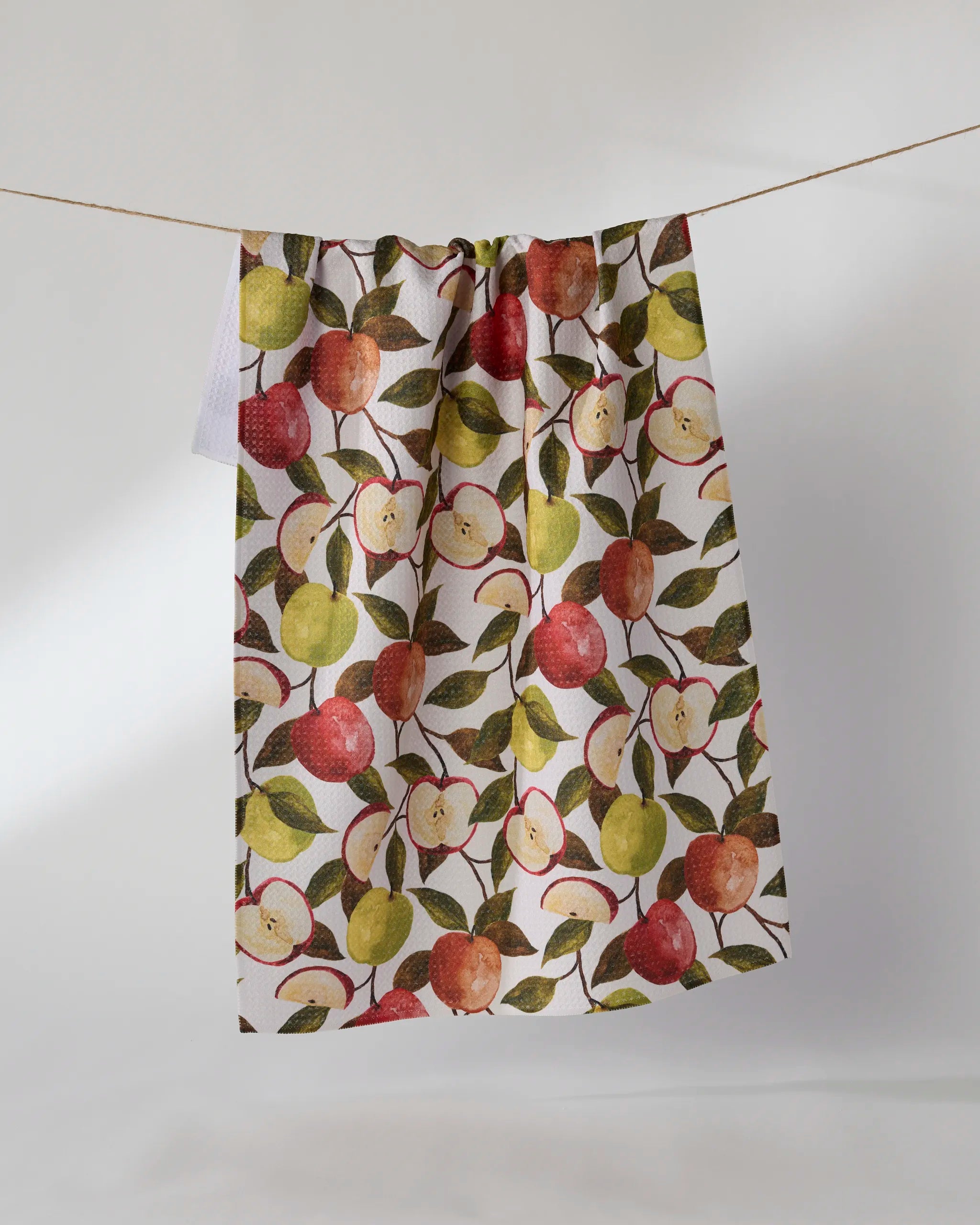 The Geometry Honeycrisp tea towel, featuring a white ultra-absorbent fabric with red and green apples and green leaves, hangs on a clothesline against a plain light background.