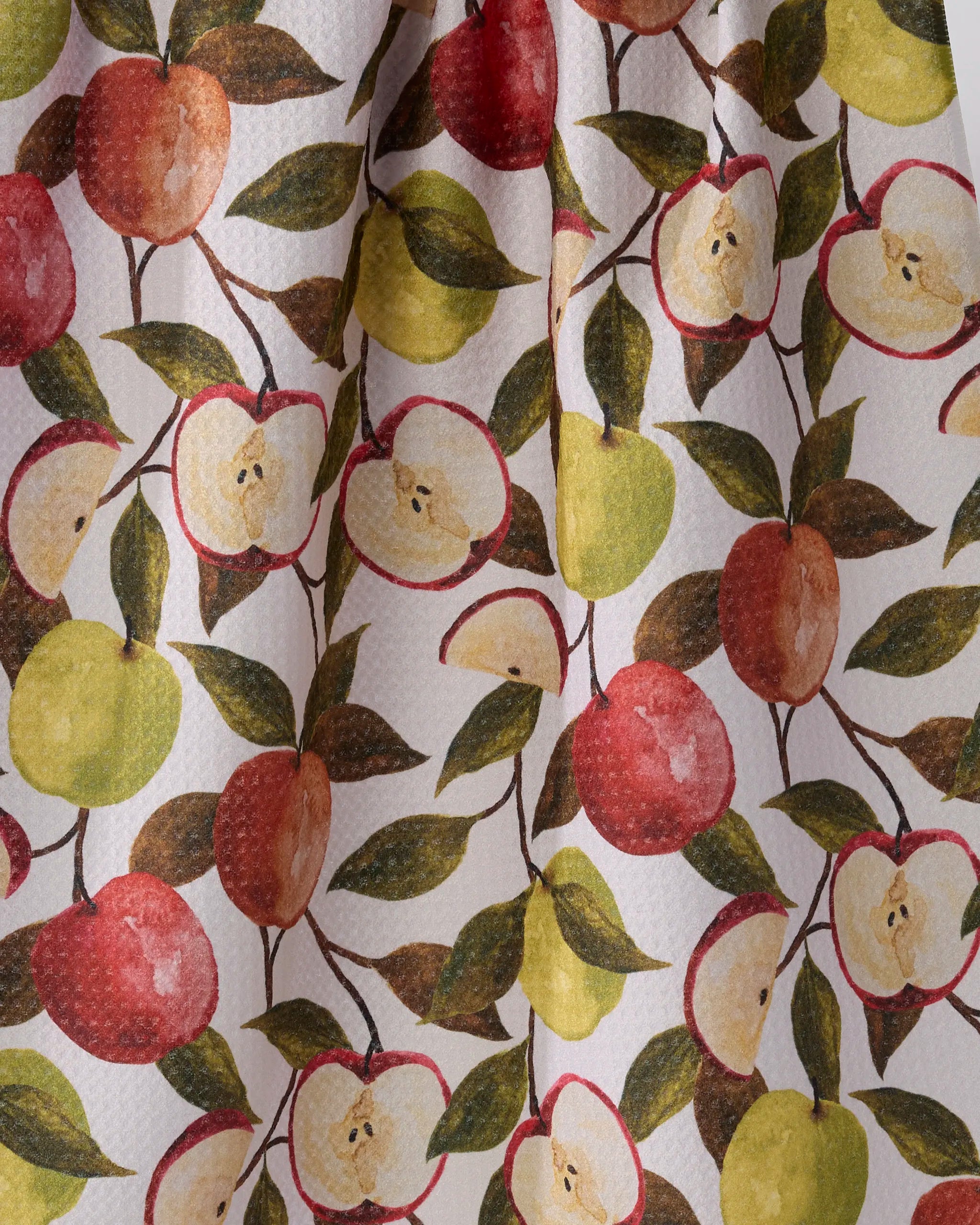 The Geometry Honeycrisp tea towel features painted red, green, and yellow apples—whole and halved with green leaves—on a textured white background.