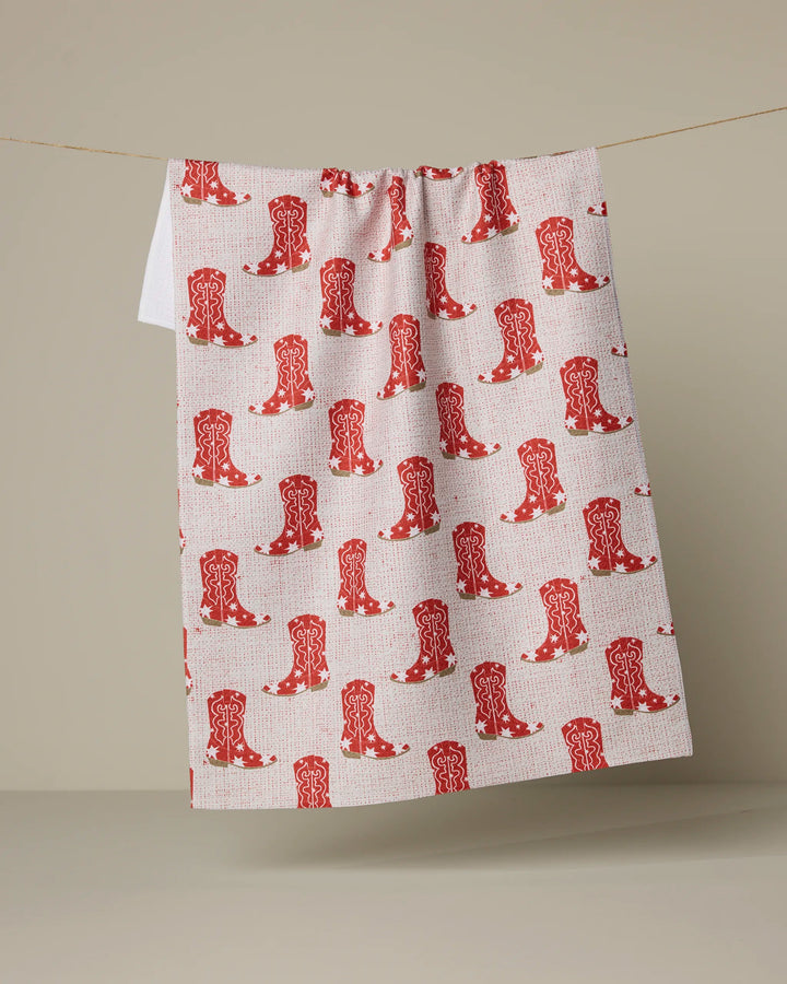 The Honky Tonk kitchen towel by Geometry features a repeating red cowboy boot pattern and hangs on a line against a neutral beige background, crafted from recycled materials.