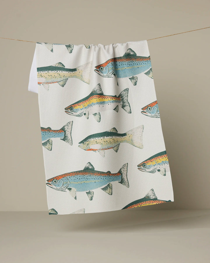 The Geometry "Hooked on Fishing" tea towel, ultra-absorbent and white with colorful blue, green, and orange fish illustrations, hangs on a clothesline against a plain beige background.