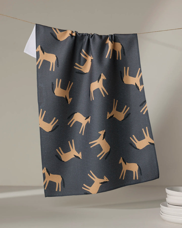 The "Horses" tea towel by Coit Creative, featuring a repeating tan horse pattern, hangs above white plates against a neutral background.