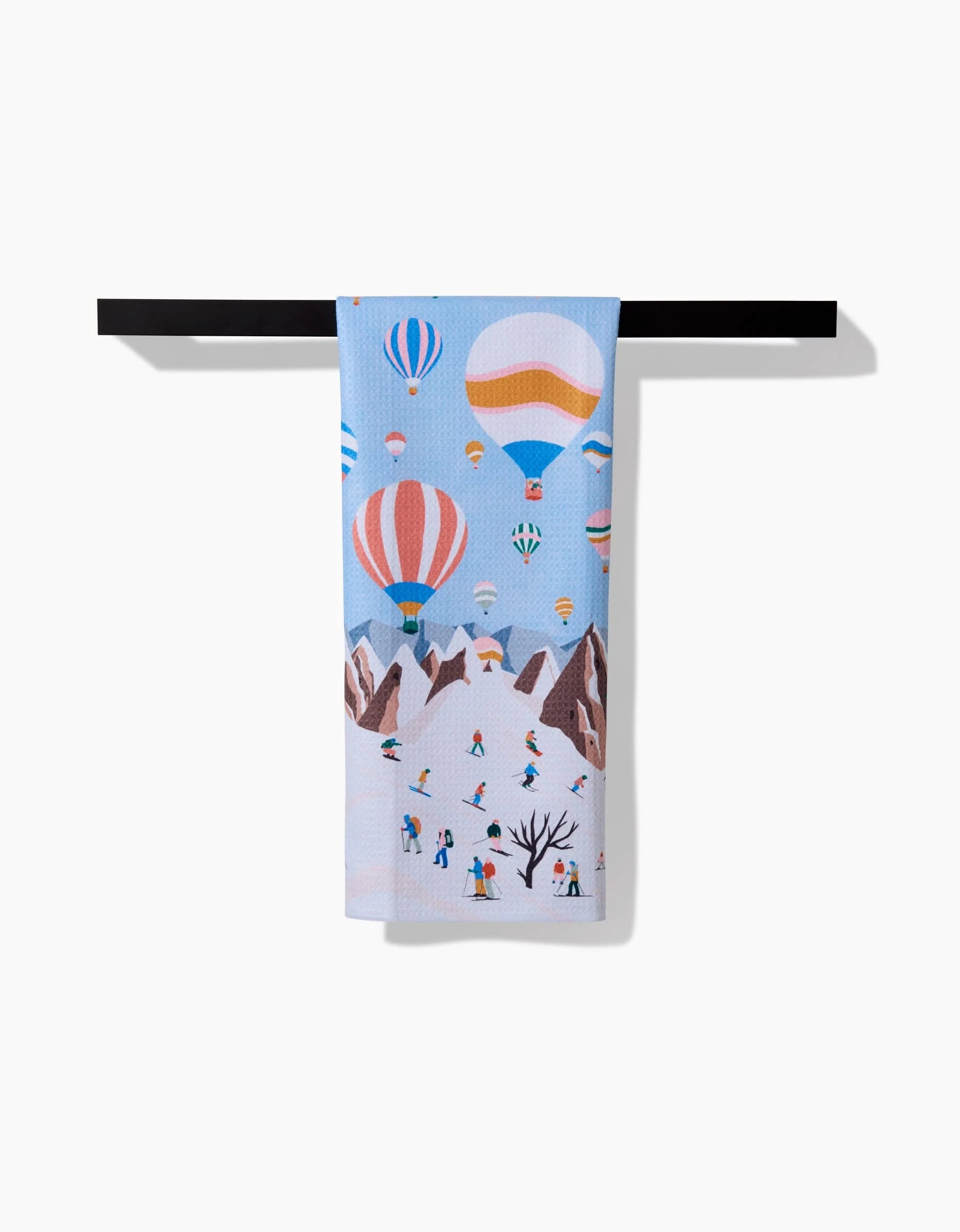 The Ceyda Alasar Hot Air Balloons tea towel hangs on a black rod, showing vibrant balloons over brown mountains, with skiers and snowboarders enjoying snowy slopes below.