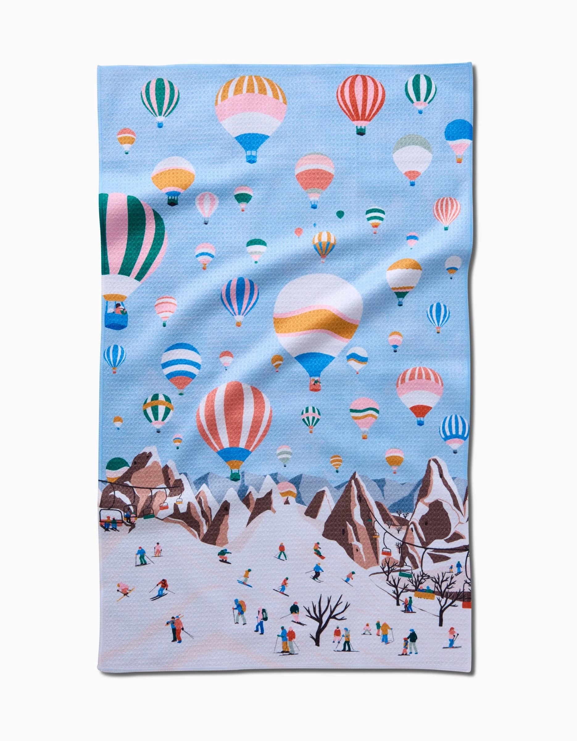 The Hot Air Balloons tea towel by Ceyda Alasar features colorful balloons in a blue sky above snowy mountains, with people skiing below—a lively design made from recycled materials.