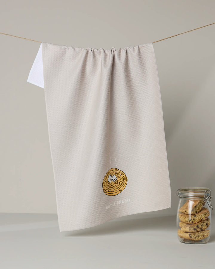The Rosa Kichline "Hot and Fresh" tea towel, embroidered with a cookie design, hangs beside a glass jar of chocolate chip cookies—adding charm to any kitchen.