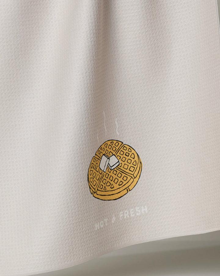 The "Hot and Fresh" tea towel by Rosa Kichline features a white textured design with a steaming waffle, a pat of butter, and the words HOT + FRESH printed below the illustration.