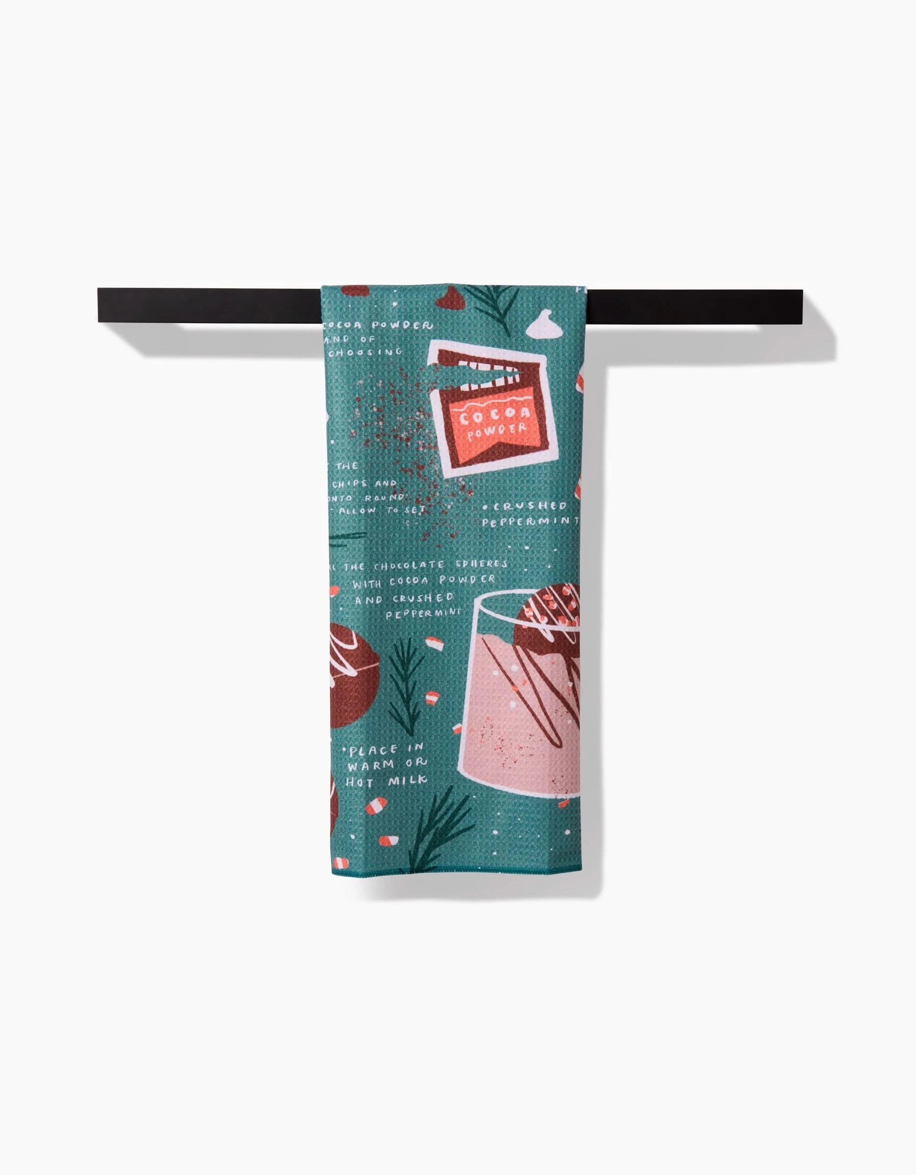 A teal Hot Cocoa Bomb kitchen towel by Janna Sue Design, featuring illustrated mugs of cocoa, cocoa powder, instructions, and decorative accents, hangs folded over a dark bar against a white background.