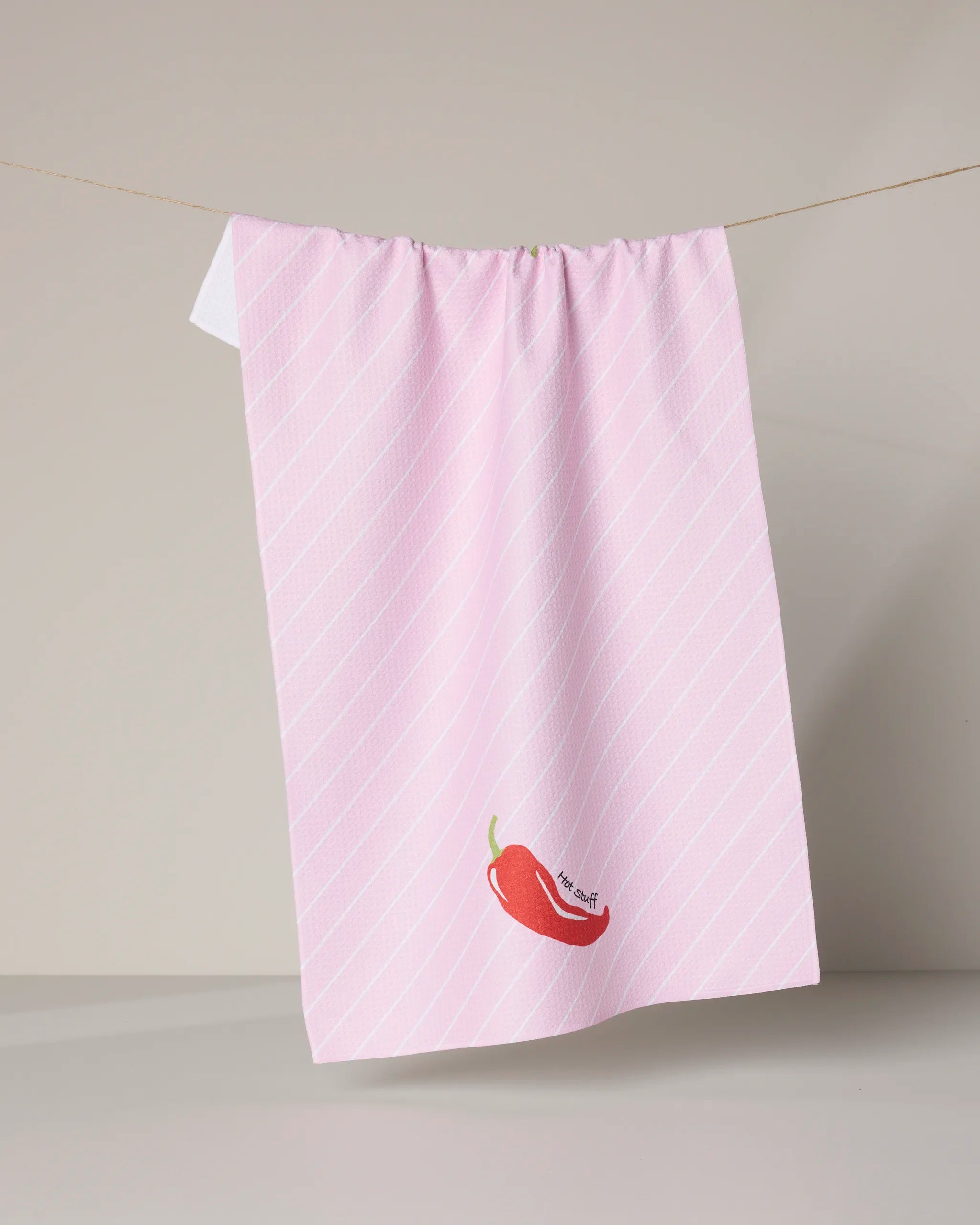 Hot Stuff by Geometry: a pink kitchen towel crafted from recycled materials, with diagonal white stripes and a red chili pepper illustration hanging on a clothesline over a neutral background.