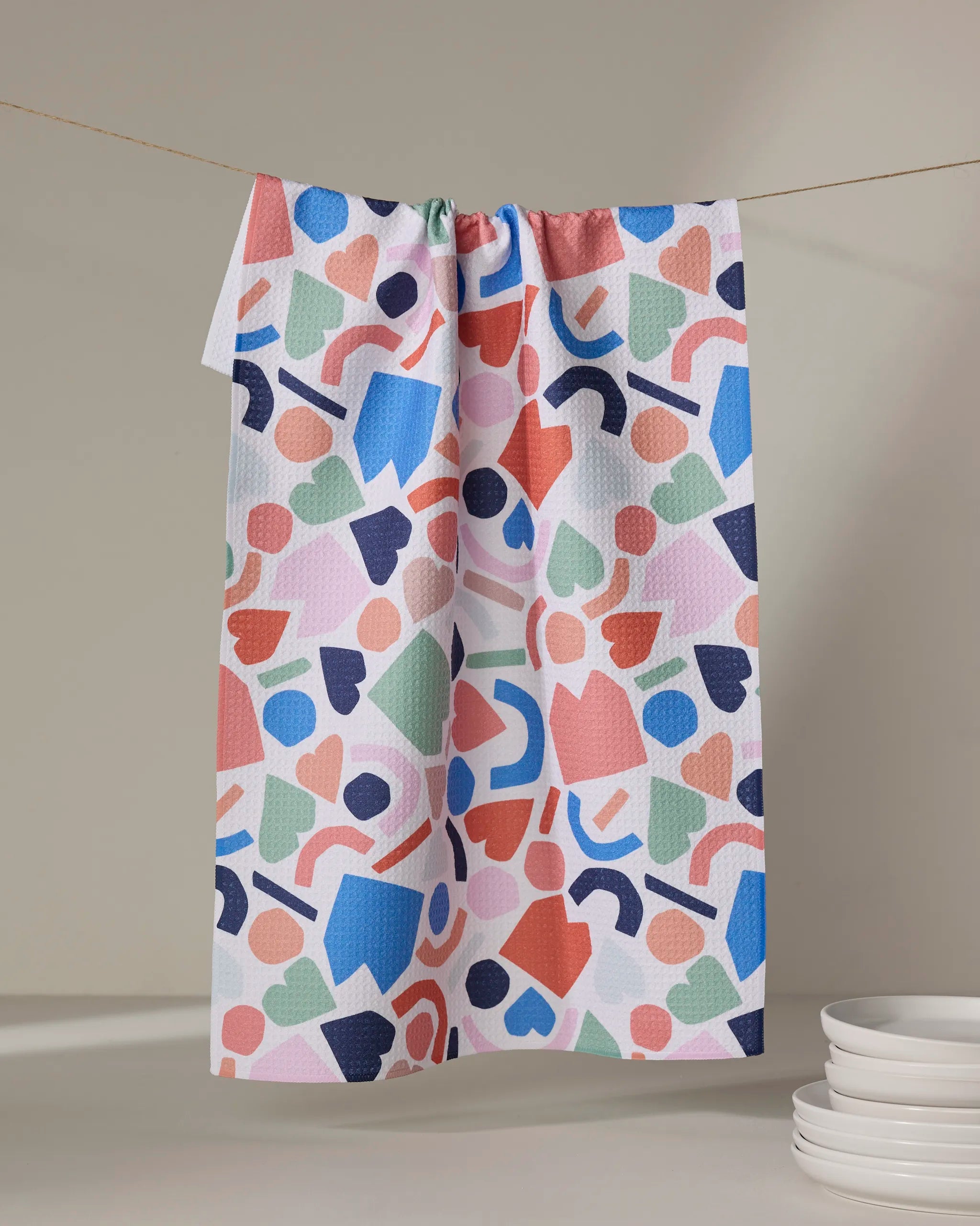 The "House Party" tea towel by Claire Iglesias, featuring colorful abstract geometric shapes, hangs on a string against a neutral background with a stack of white plates nearby.