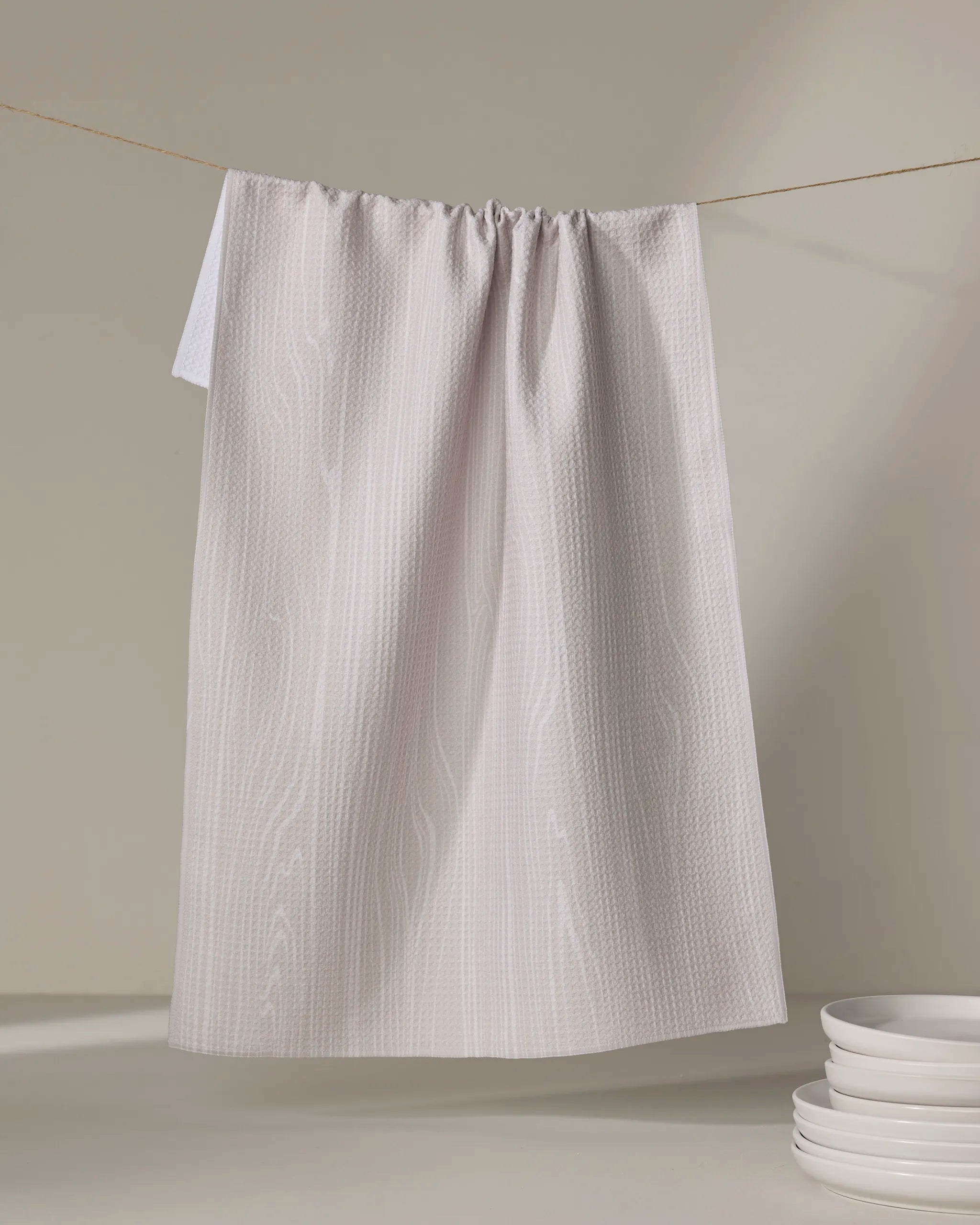The Houston Wood tea towel by Kailee Wright, featuring a light gray textured pattern and made from recycled materials, hangs on a string with a stack of white plates in the lower right corner against a neutral background.
