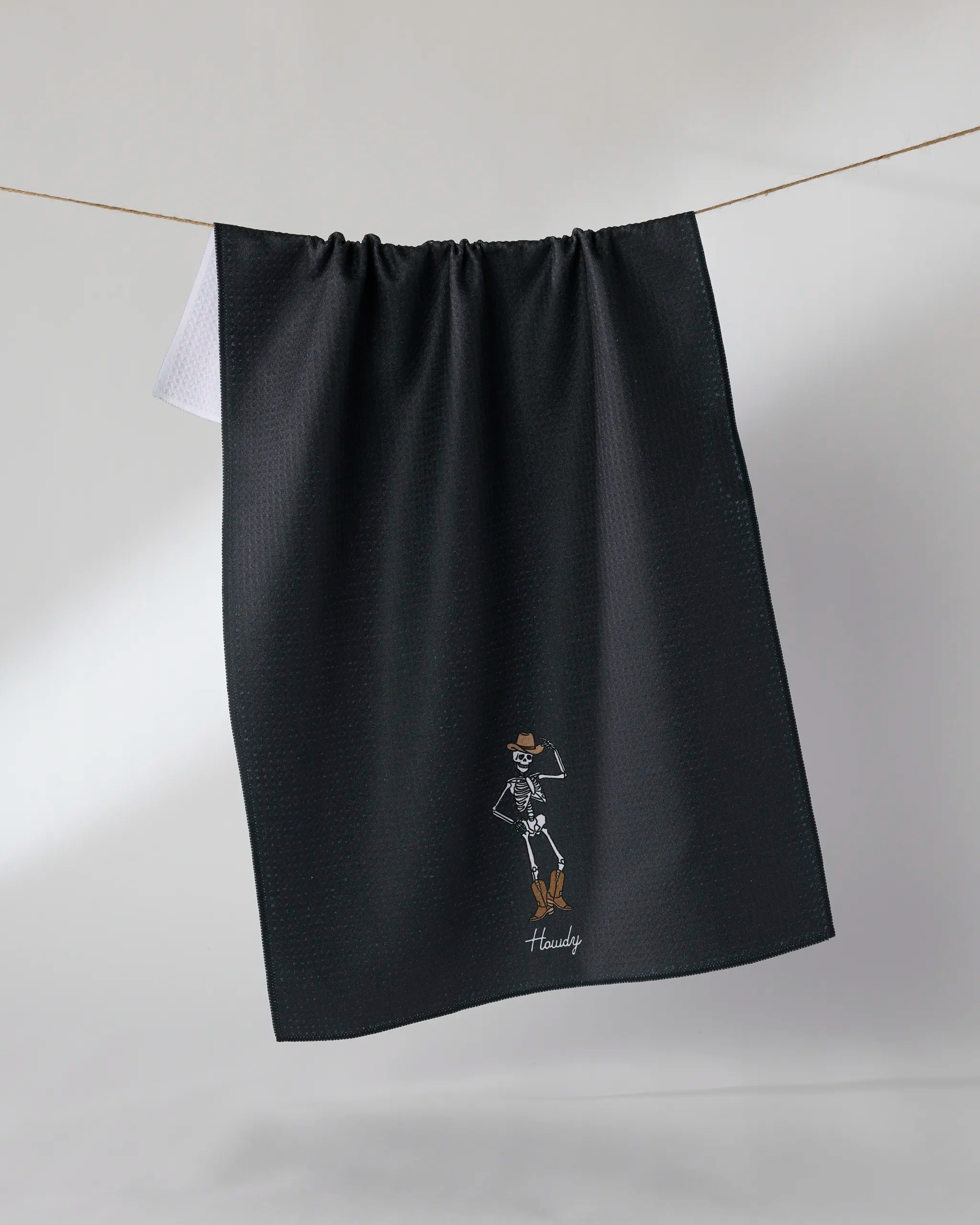 The Geometry Howdy Cassondra Black kitchen towel, made from recycled materials, features a small embroidered cowboy skeleton in boots and hat near the bottom with "Howdy" underneath, hanging on a line. Eco-friendly and stylish.