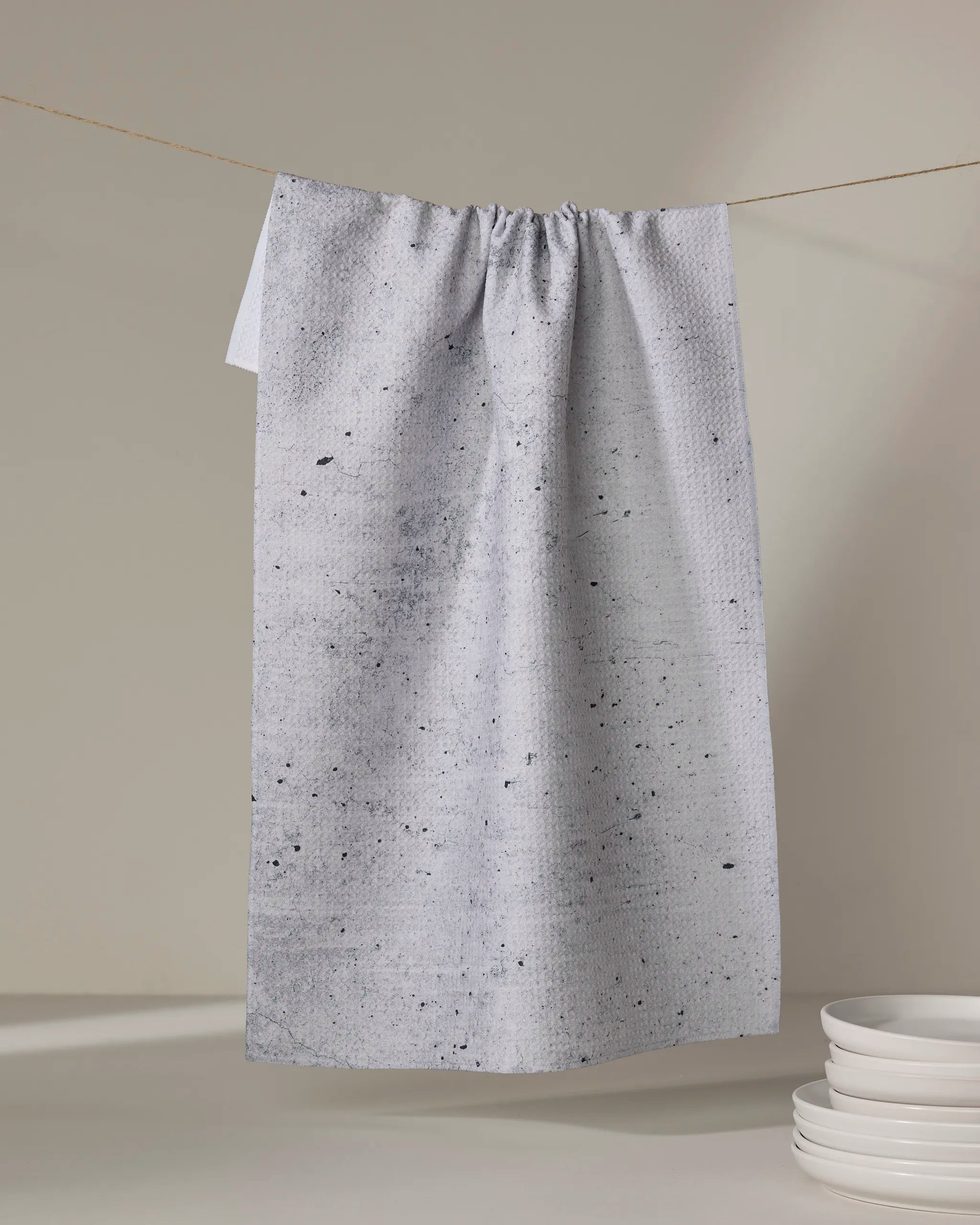 The Kailee Wright Hudson Grey kitchen tea towel, featuring a light gray speckled design, hangs on a clothesline against a neutral backdrop with a stack of white plates resting below.