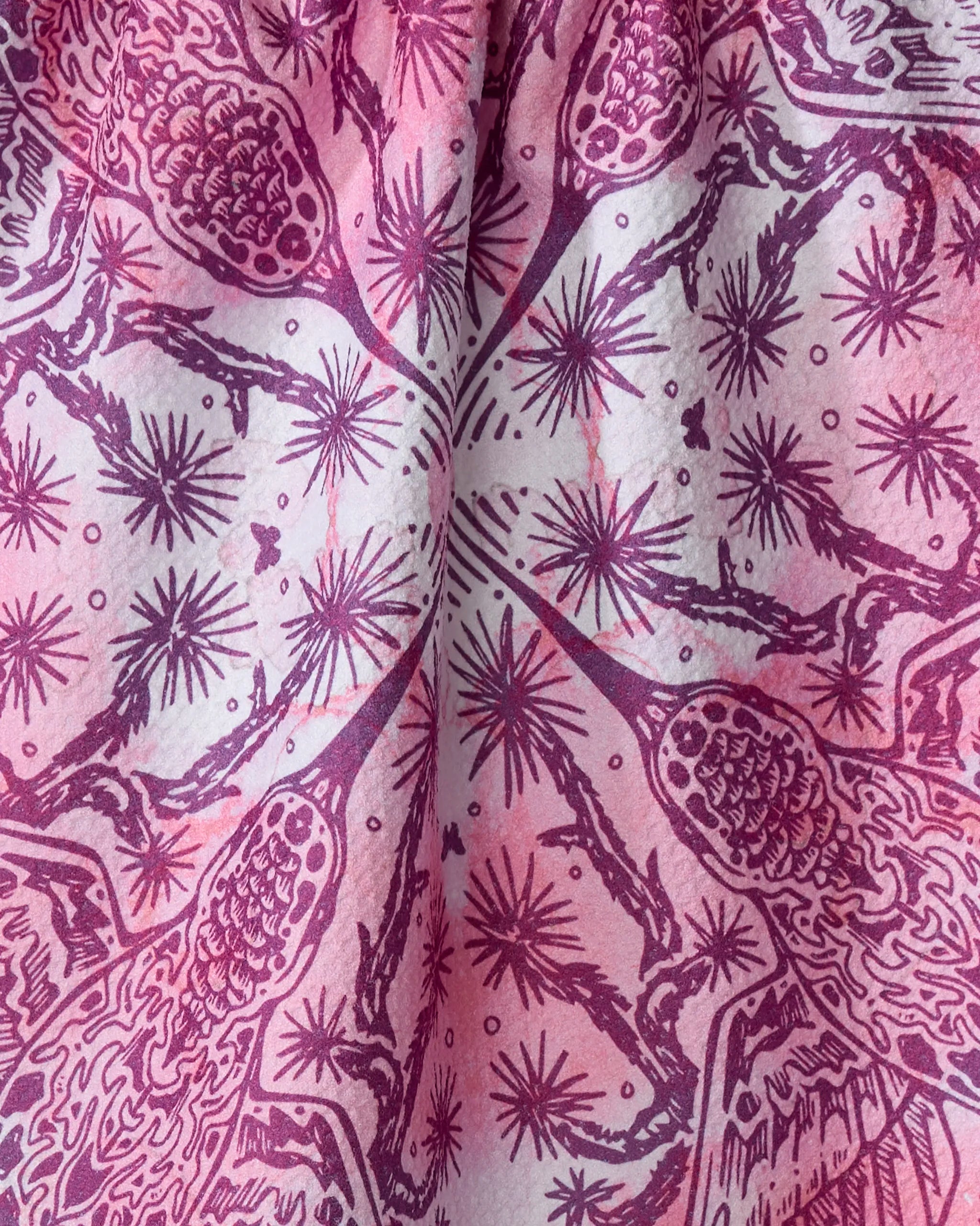 The Skye Walker Hummingbird tea towel features a pink and purple pattern with symmetrical narwhals and sea plants radiating from the center. Absorbent fabric folds create textured shadows, making it perfect for brightening your kitchen.
