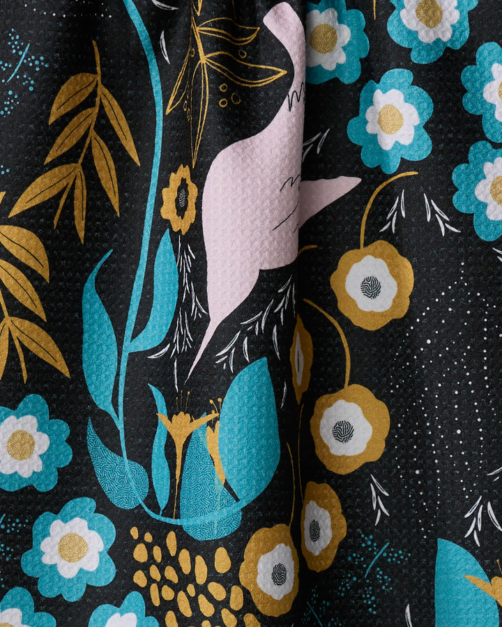 A close-up of the Julianne Haness Hummingbird Petals tea towel, showcasing its textured Geoweave fabric with teal, gold, white, and beige floral patterns on a black background and subtle folds.