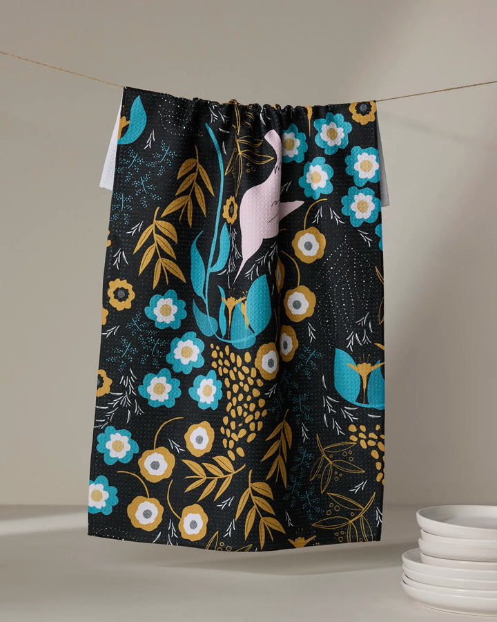 The Hummingbird Petals tea towel by Julianne Haness, made from recycled materials, showcases a blue, yellow, white, and pink floral and bird design. It hangs above a stack of white plates against a dark background.