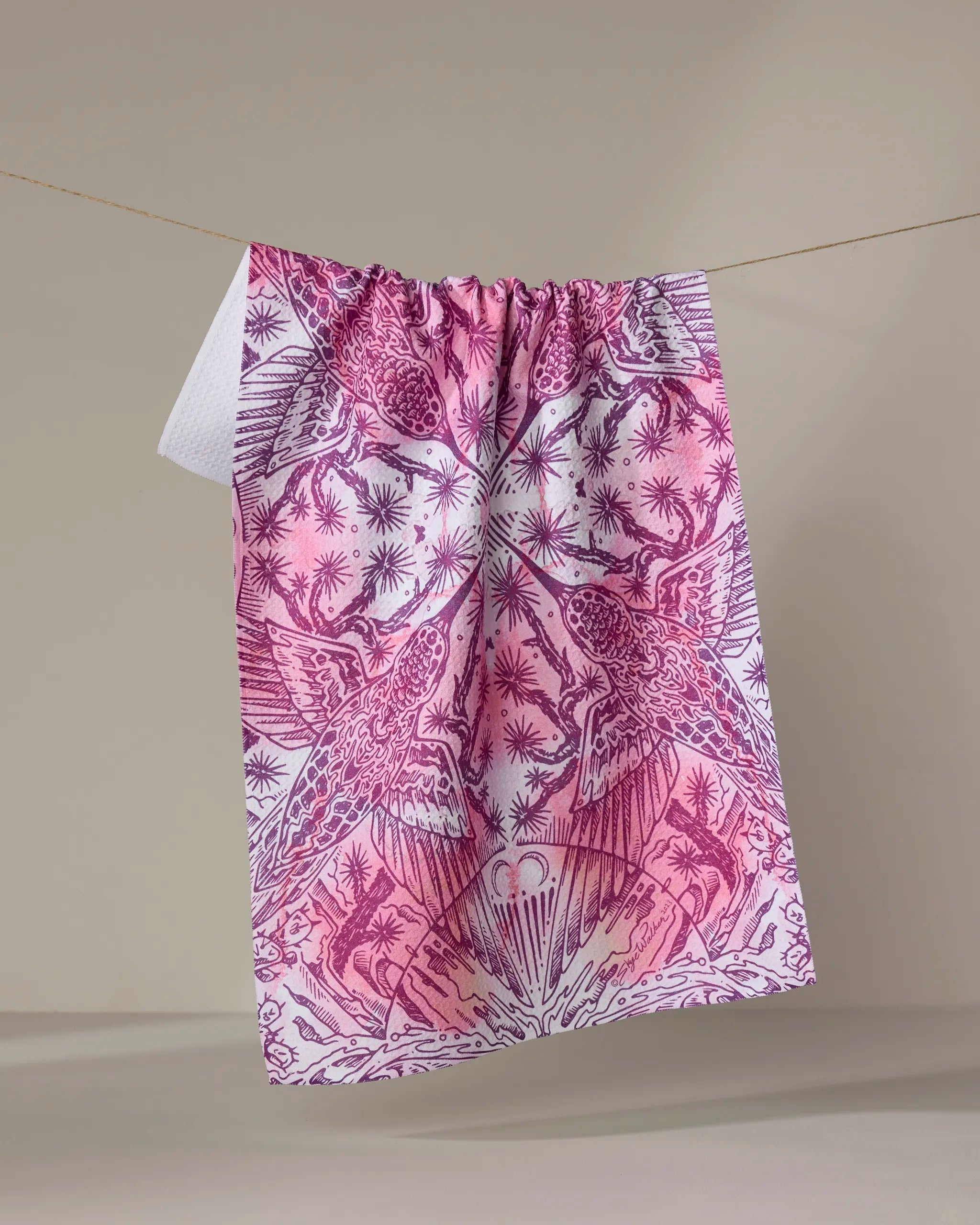 The Skye Walker "Hummingbird" tea towel, featuring pink and white patterns of birds, plants, and stars, hangs on a clothesline against a neutral background.