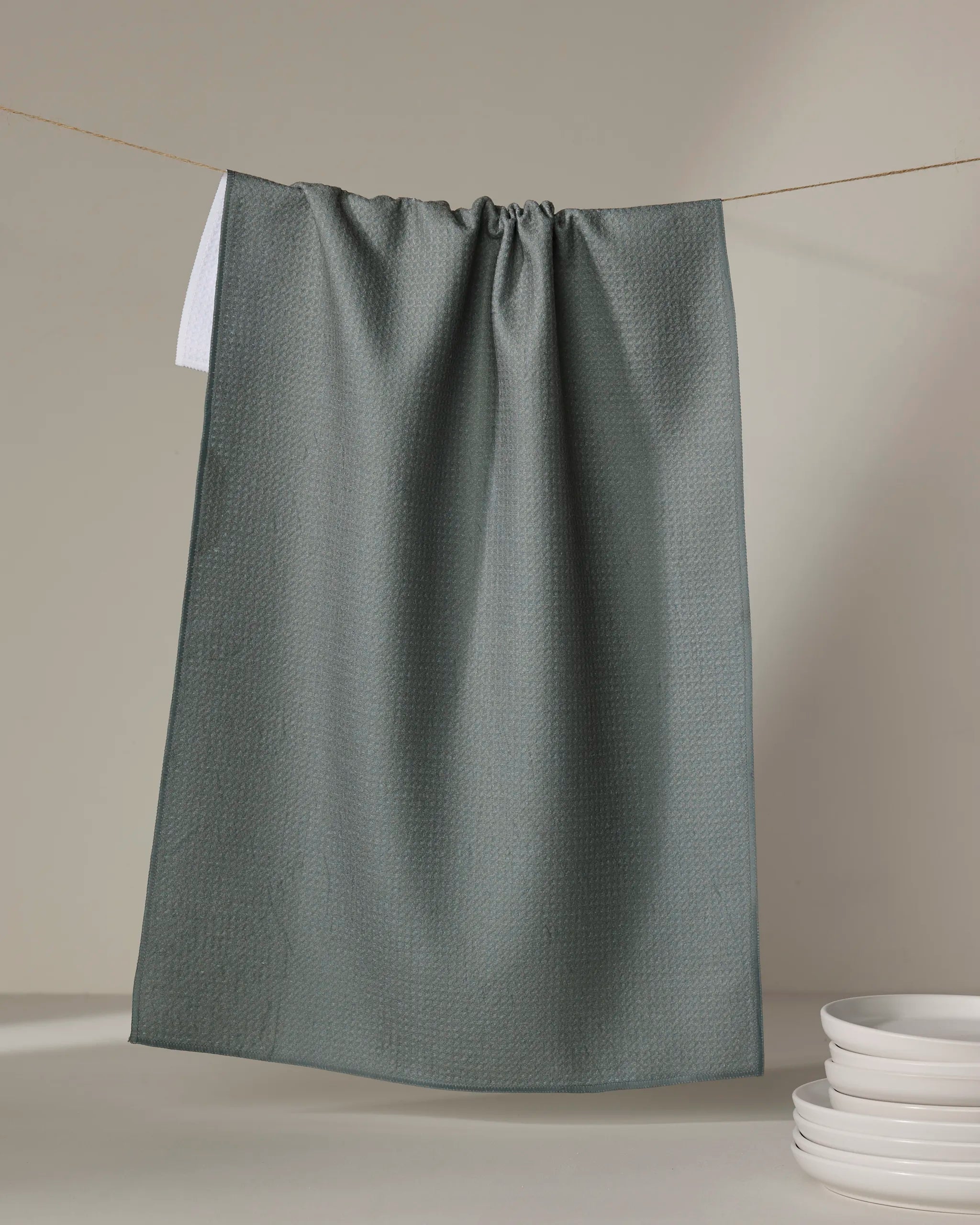 A Kailee Wright Hunter Green kitchen tea towel, made from recycled materials, hangs on a string against a neutral background, with a stack of white plates on the lower right side.