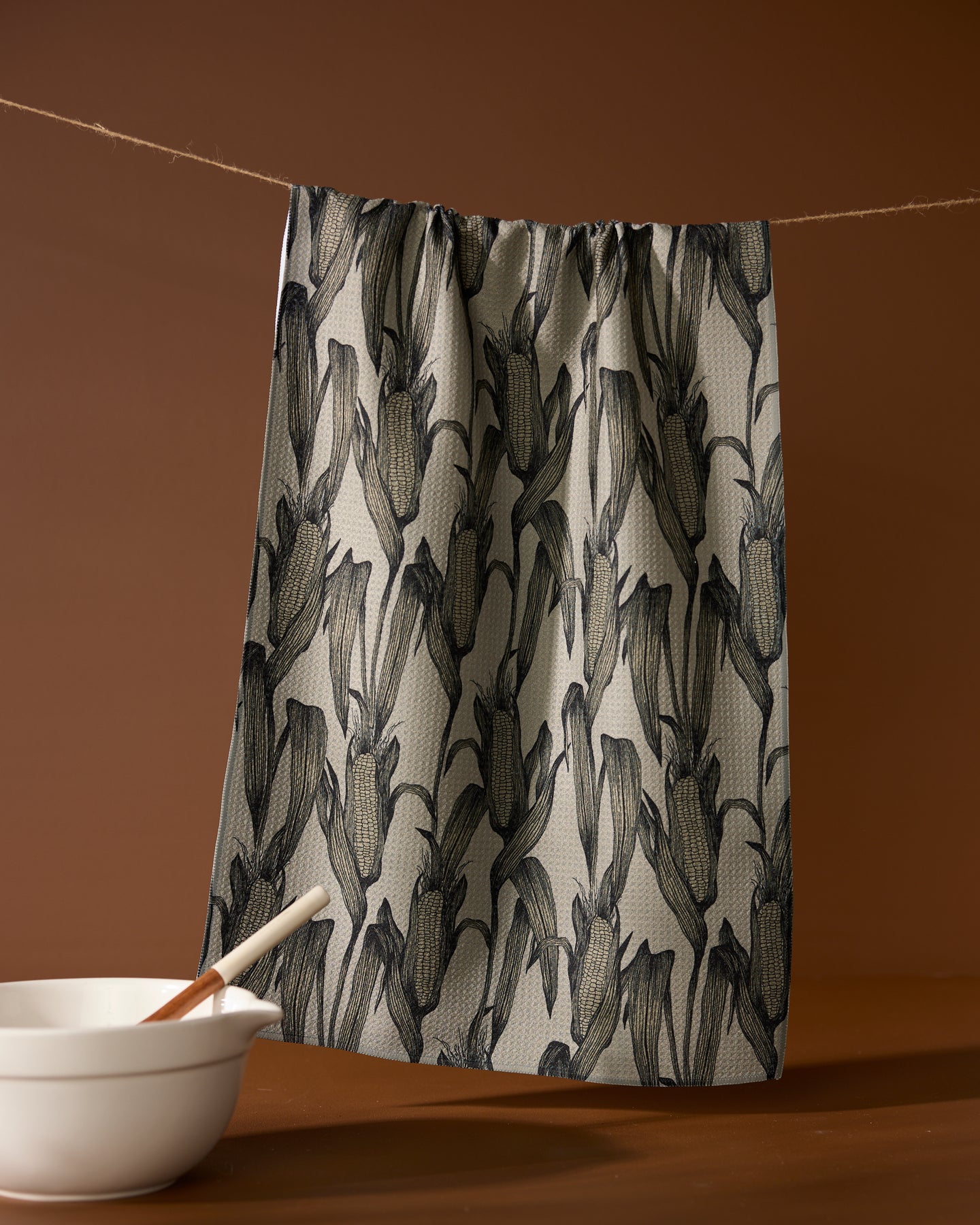 The Geometry Husk recycled tea towel, featuring a black botanical pattern, hangs on a string against a brown backdrop. A white bowl with a wooden spoon sits in front, enhancing the inviting kitchen scene.