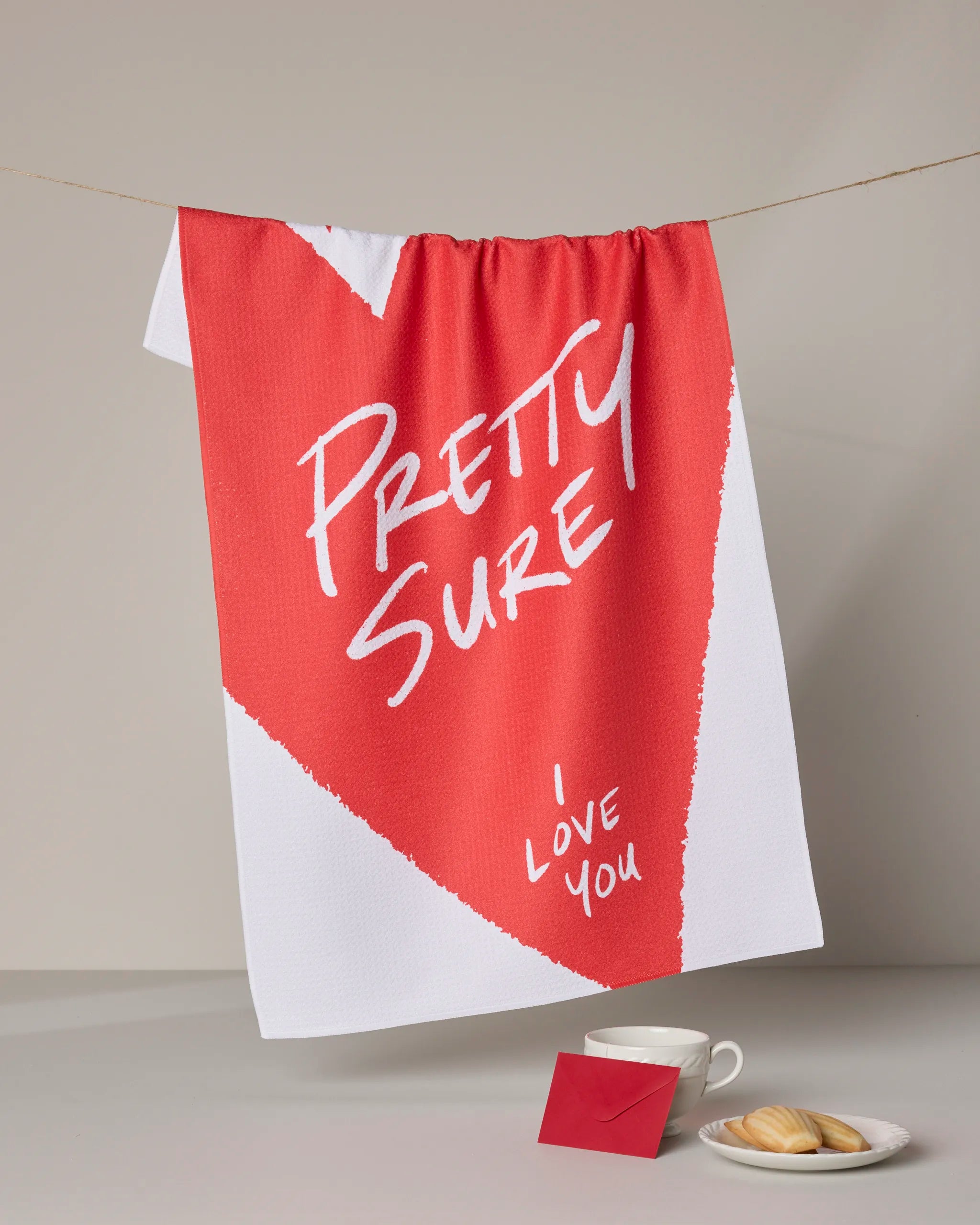 A red and white Geometry "I Love Thee" kitchen towel hangs on a line. Below, a red envelope, white cup and saucer, and cookies on a plate complete this charming scene.