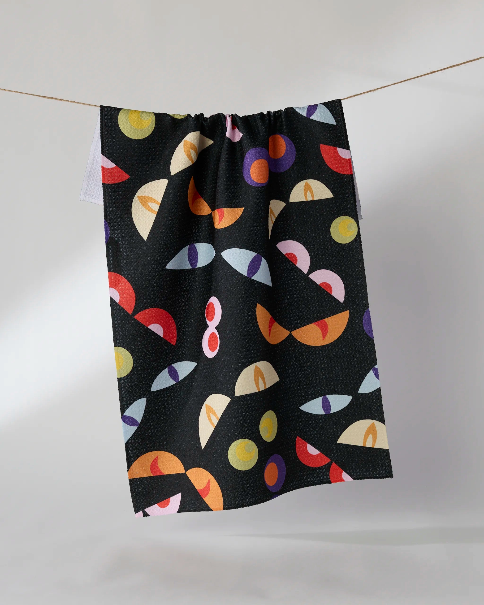The Geometry "I See You" tea towel, made from black Geoweave fabric and featuring a colorful abstract pattern of eye-like shapes, half-moons, and circles in yellow, red, purple, and orange, hangs on a clothesline.