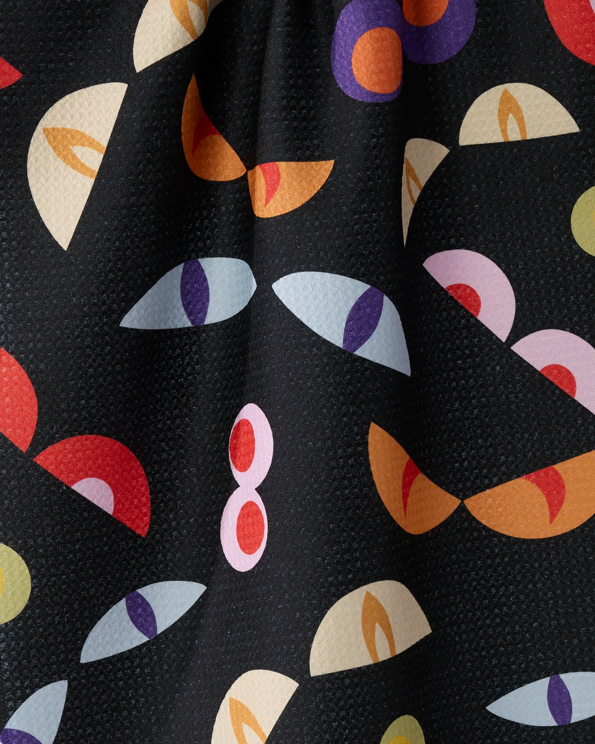 Geometry’s “I See You” kitchen tea towel features colorful abstract shapes like stylized eyes and semi-circles in red, purple, yellow, and pink scattered on a textured black background.