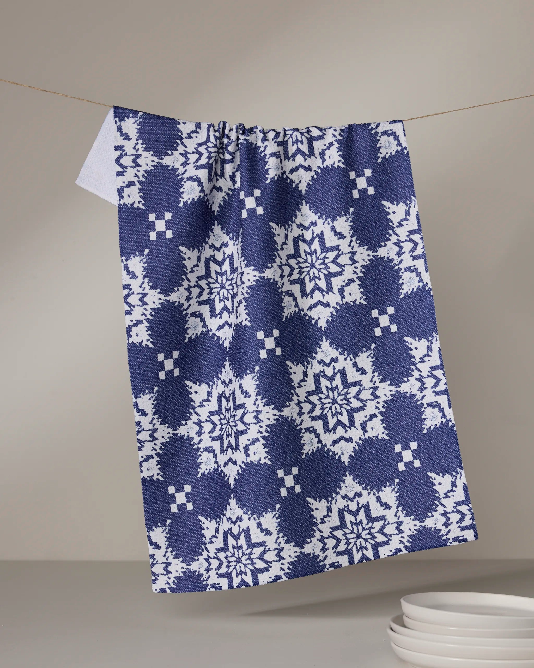 The Geometry Iceflower Blue kitchen towel, featuring a blue and white geometric snowflake pattern and made from recycled materials, hangs on a line with stacked white plates in the lower right against a neutral backdrop.
