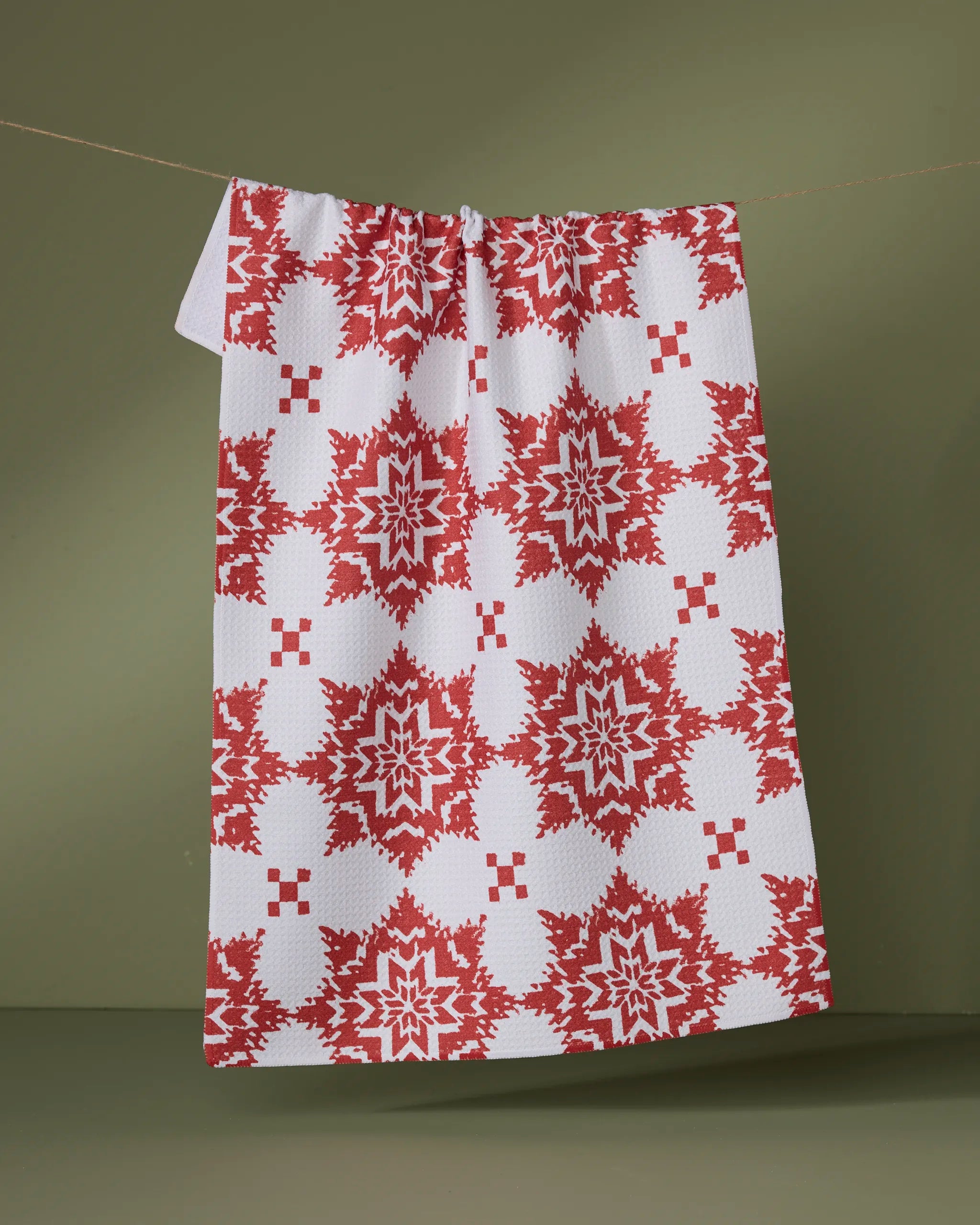 The Geometry Iceflower tea towel features a red and white geometric, snowflake-inspired design. Made from recycled materials, it brings style and sustainability to your kitchen décor. Shown hanging against an olive green background.