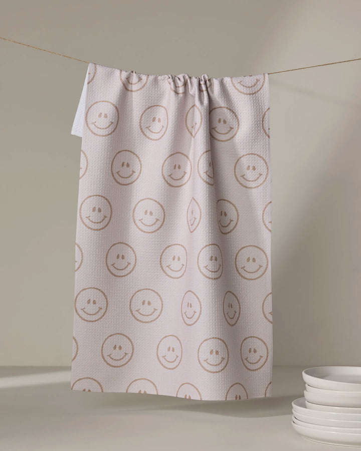 The "I'm Blushing" tea towel by Kelsi Fullmer, featuring cheerful smiley faces, hangs on a line to dry. A small stack of white plates sits in the lower right corner against a neutral background.