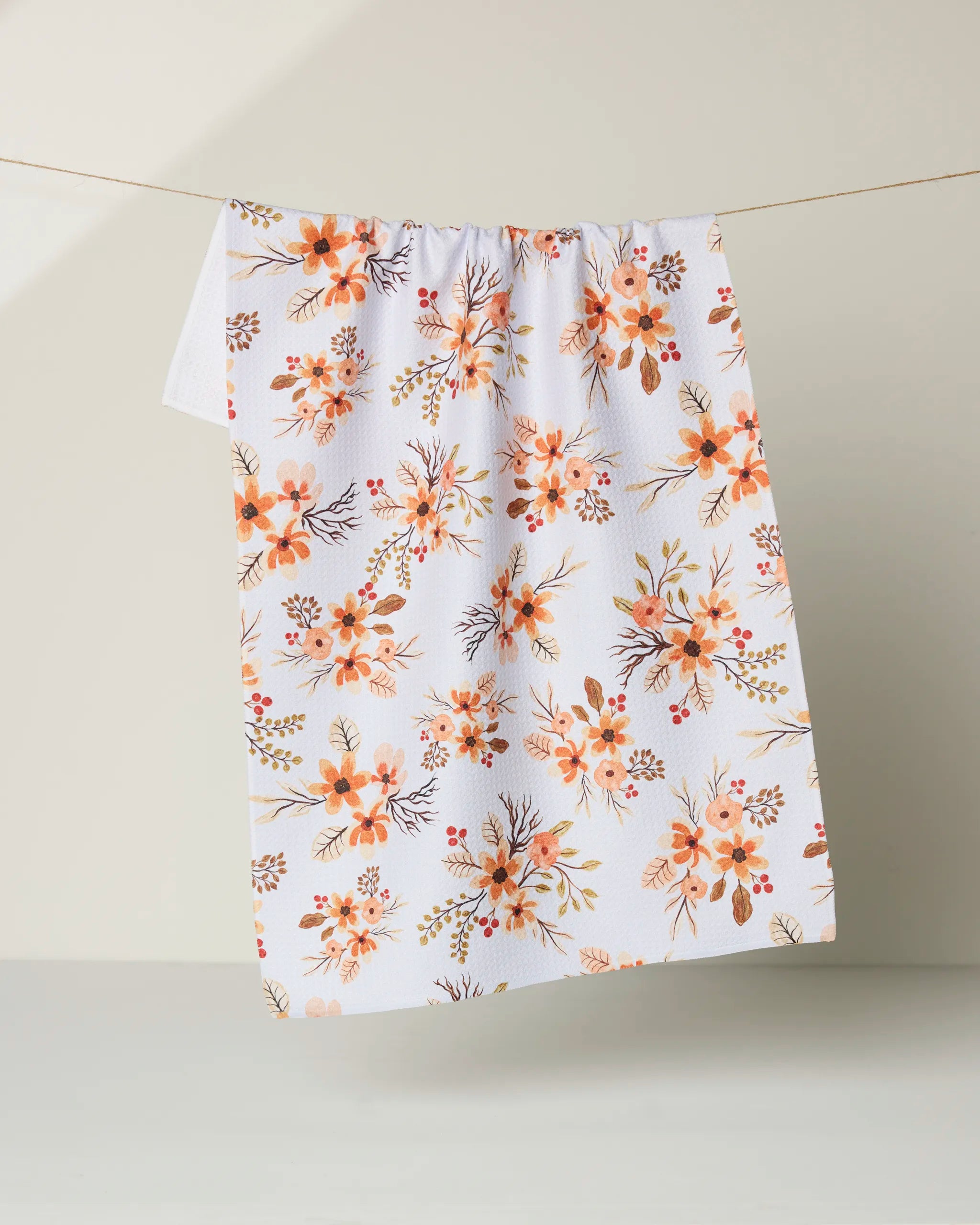 The "In Bloom" tea towel by Vaishali Design Studio, featuring an orange and yellow floral pattern, hangs on a string against a neutral background.