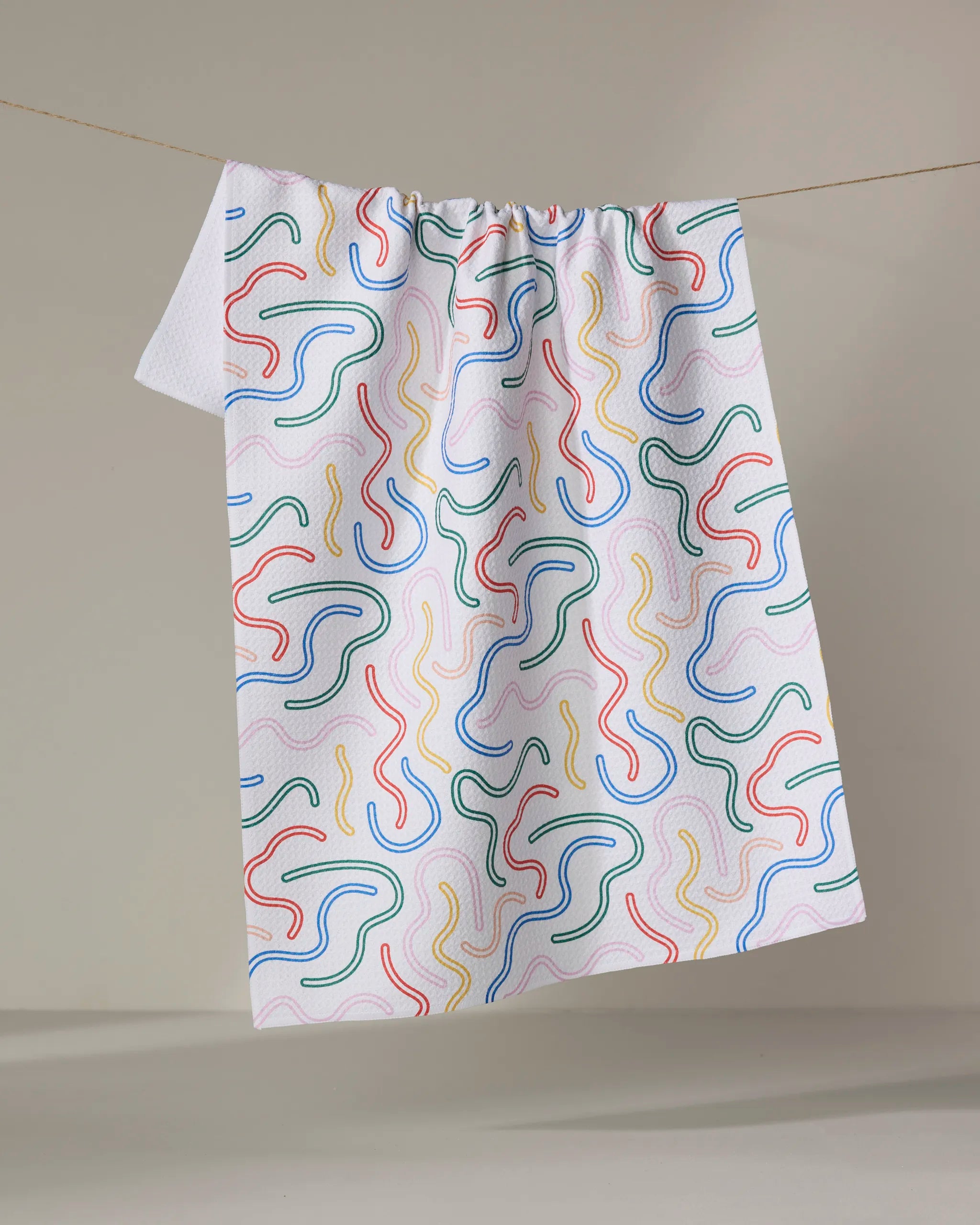 The "In the Moment" tea towel by Happy Hemera features a white textured fabric with colorful squiggly lines in blue, red, yellow, and green, hanging on a clothesline against a neutral backdrop.
