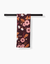 The Ashley Celena In The Oaks recycled kitchen towel in dark maroon with pink, white, and orange floral pattern is neatly draped over a black bar against a white background.