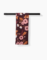 The Ashley Celena In The Oaks recycled kitchen towel in dark maroon with pink, white, and orange floral pattern is neatly draped over a black bar against a white background.