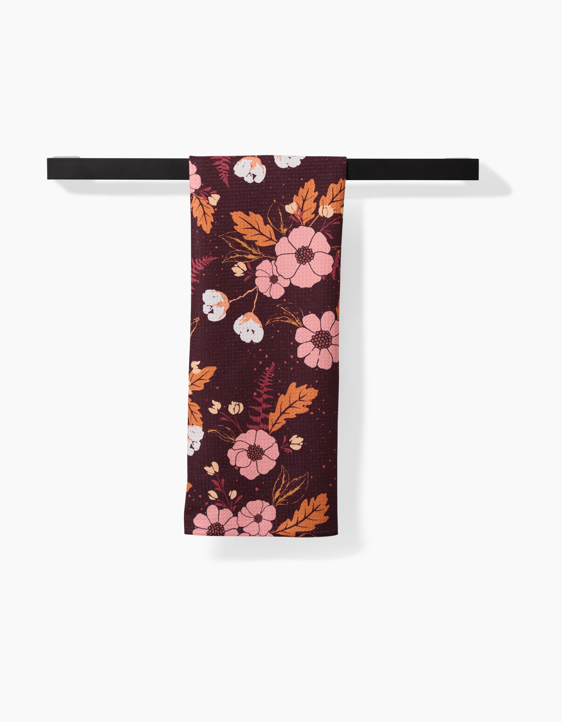 The Ashley Celena In The Oaks recycled kitchen towel in dark maroon with pink, white, and orange floral pattern is neatly draped over a black bar against a white background.