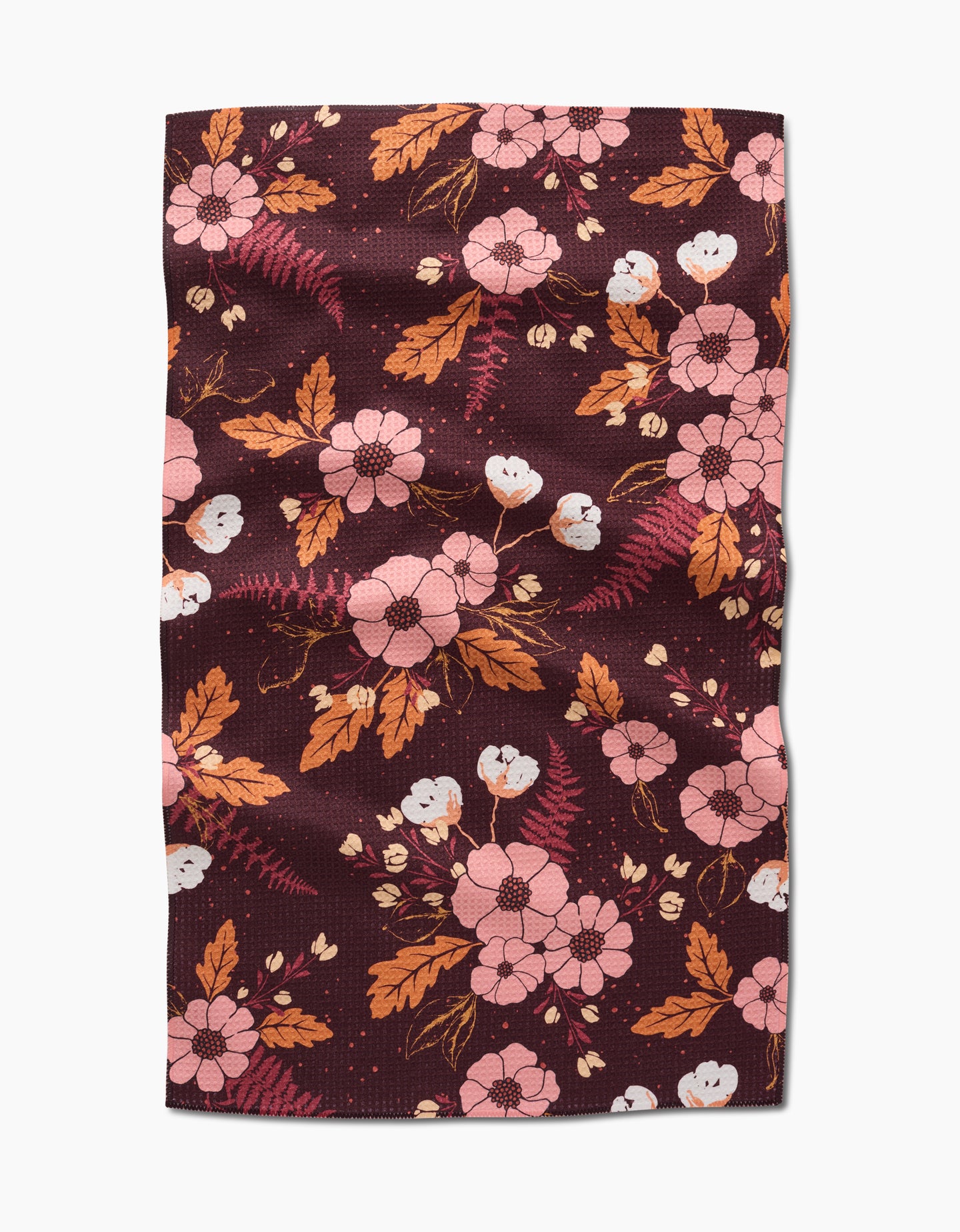 The In The Oaks tea towel by Ashley Celena features a maroon background with a floral pattern of pink flowers, orange and brown leaves, and small white buds. The rectangular fabric has a slightly wrinkled appearance.