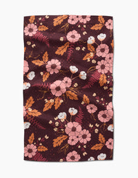 The In The Oaks tea towel by Ashley Celena features a maroon background with a floral pattern of pink flowers, orange and brown leaves, and small white buds. The rectangular fabric has a slightly wrinkled appearance.