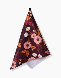 The In The Oaks kitchen towel by Ashley Celena is triangular and maroon, featuring a pink, orange, and white floral pattern. It hangs from a black loop against a white background.