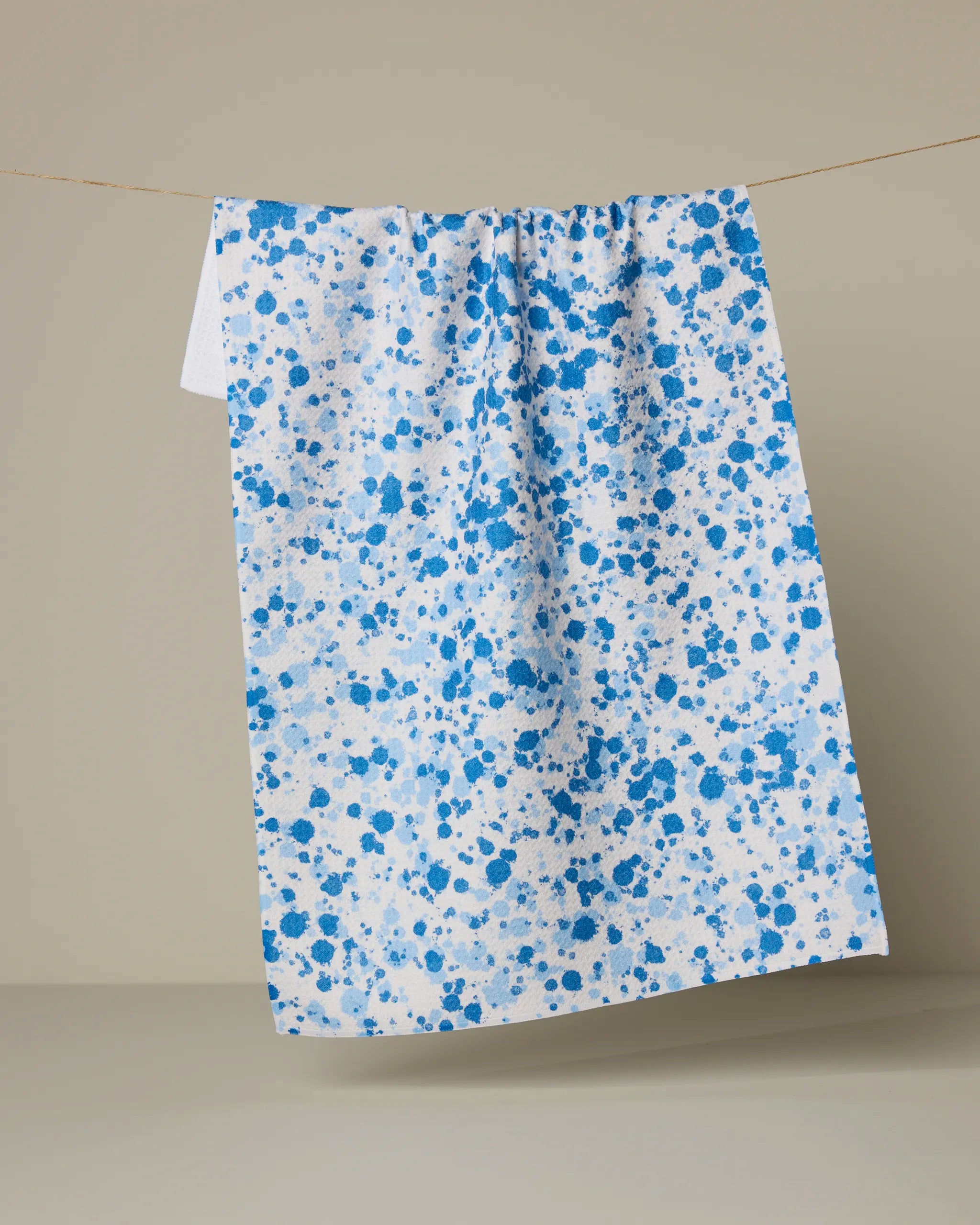 The Indigo Splash tea towel by Geometry, crafted from recycled materials with a blue paint splatter design, hangs on a clothesline against a plain beige background.